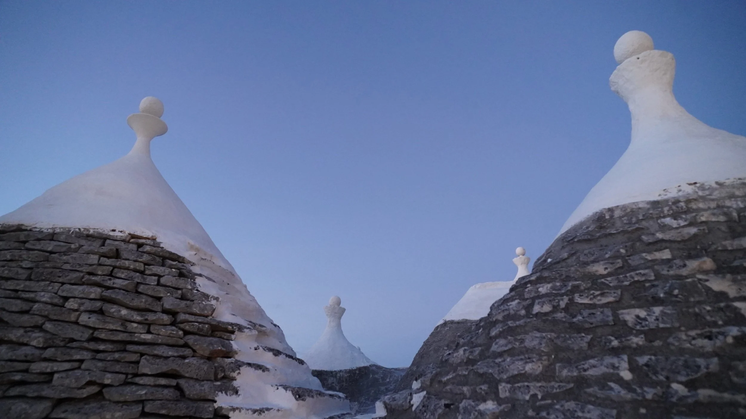 Tre coni di tetto di case con rifiniture bianche e pietre grigie, sormontati da decorativi sferici, sotto un cielo blu.