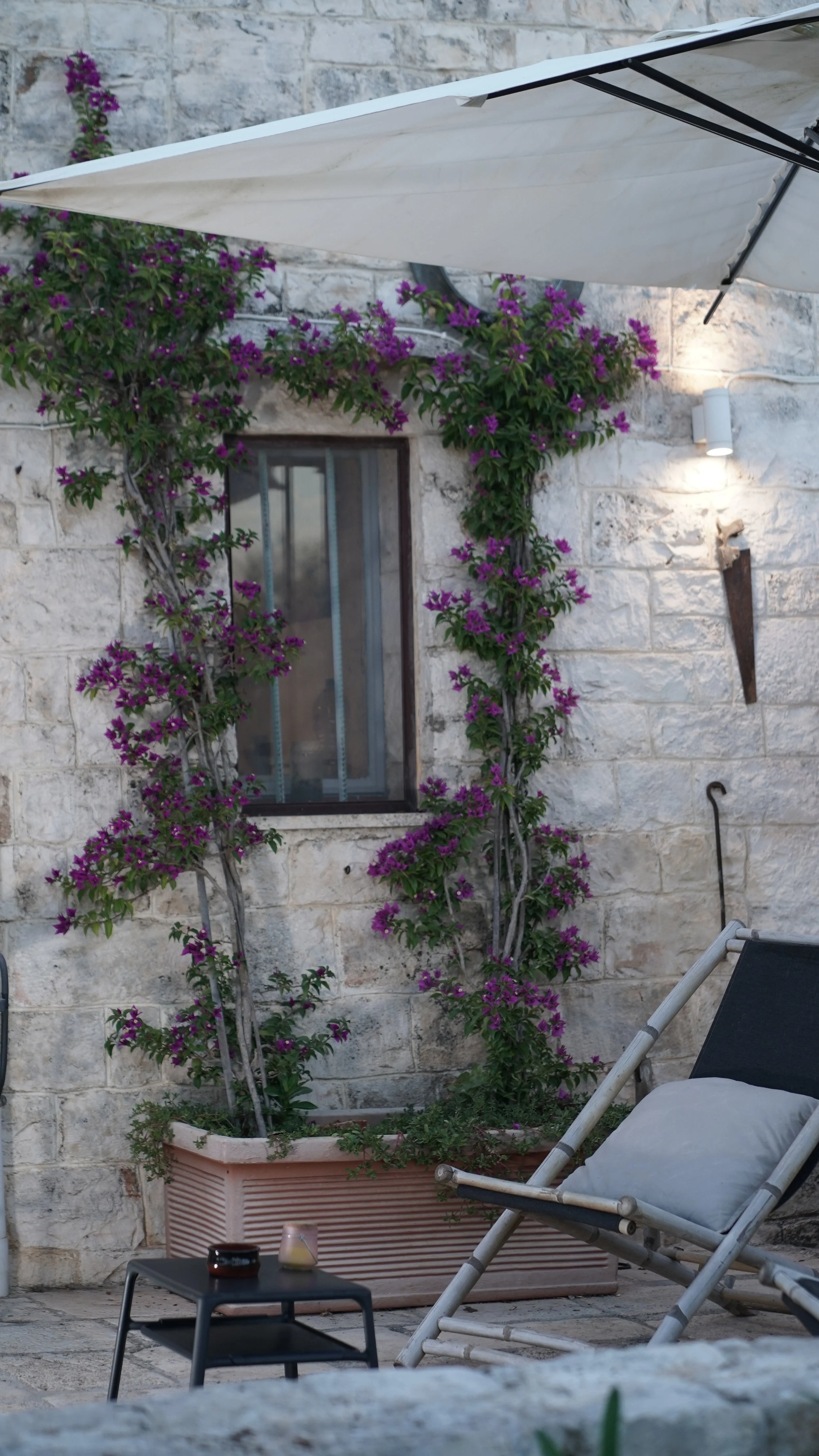 Una parete in pietra con una finestra incorniciata di viti di bouganville con fiori viola, un'ombra di ombrellone bianco, una sedia a sdraio con cuscino e un tavolo con candele o bicchieri.