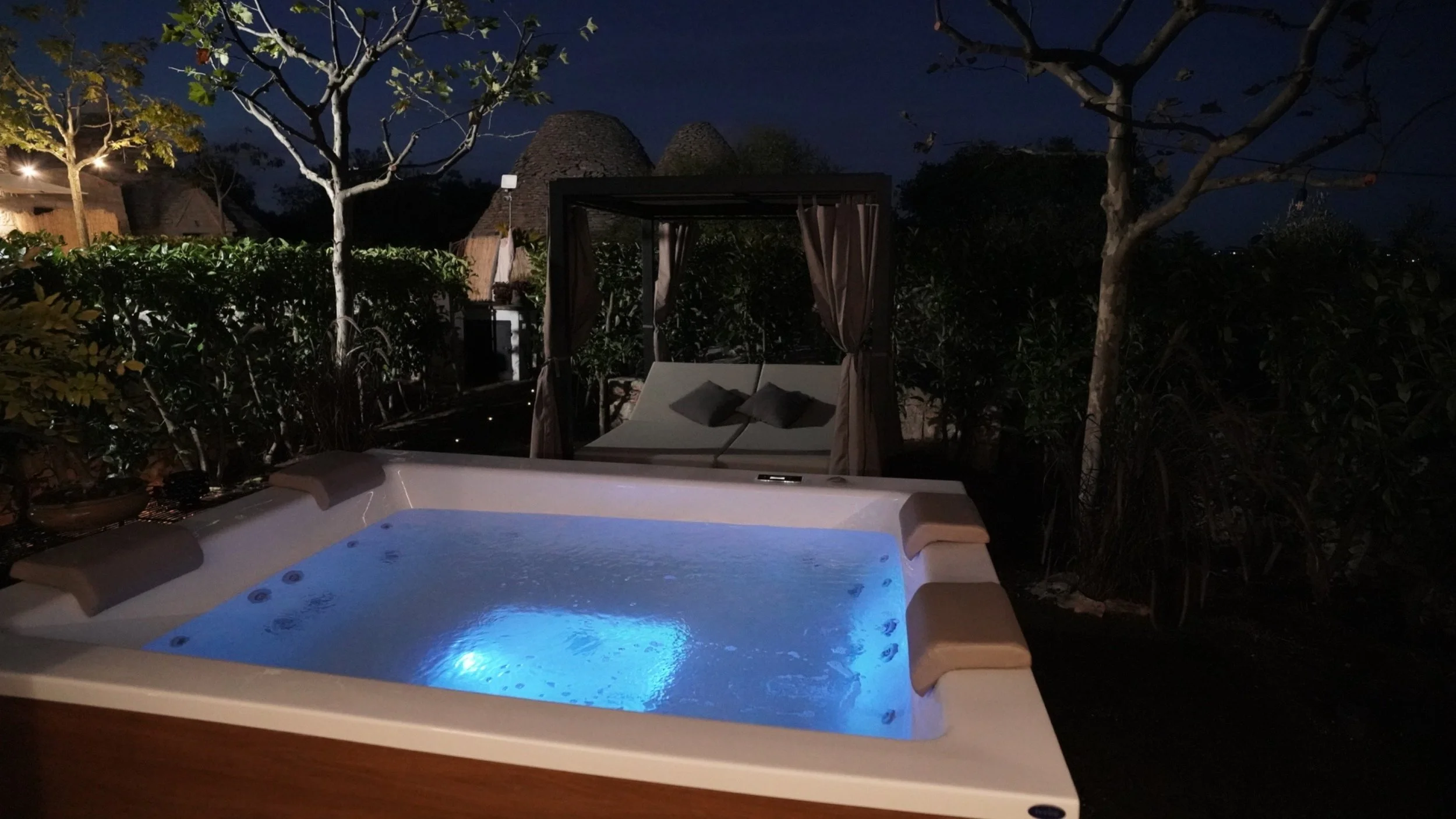 Jacuzzi illuminato di notte con due sedie a sdraio e un baldacchino sopra, circondato da alberi e vegetazione, con abitazioni di paglia sullo sfondo.
