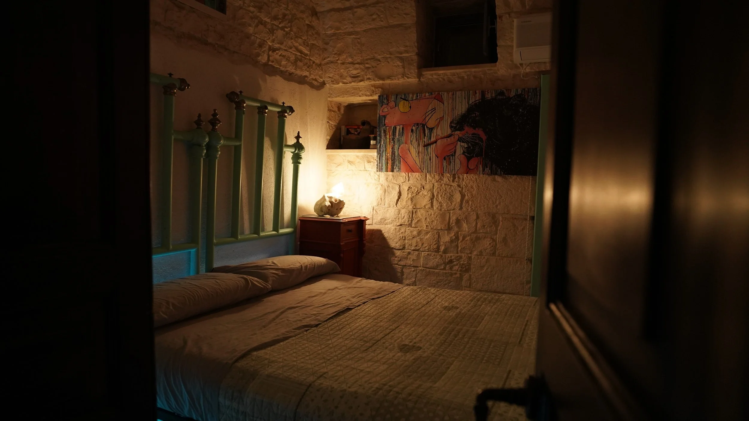 Camera da letto con pareti di pietra, letto con coperta beige, lampada accesa su comodino di legno con oggetto decorativo, quadro colorato su parete, finestra alta, aria condizionata