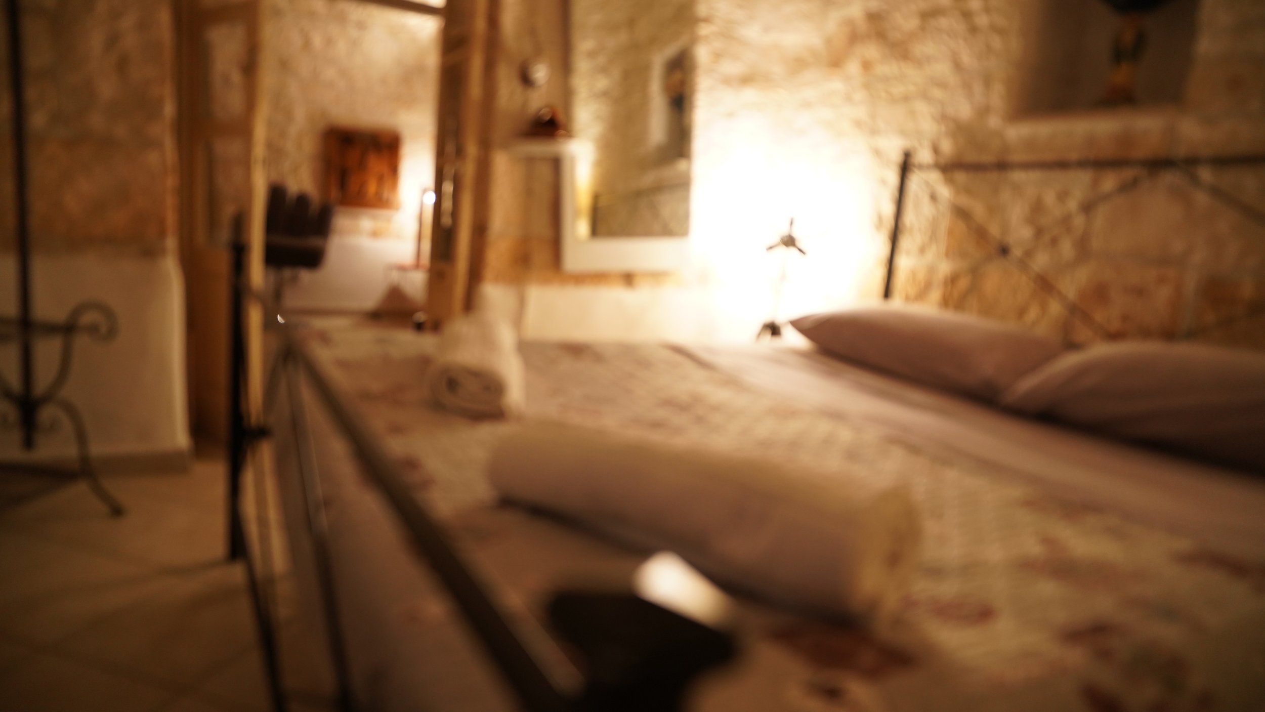 Una stanza da letto con un letto con biancheria colorata, un specchio appeso alla parete e una lampada accesa. La stanza ha pareti di pietra e un arredamento rustico, con tende e complementi in stile vintage.