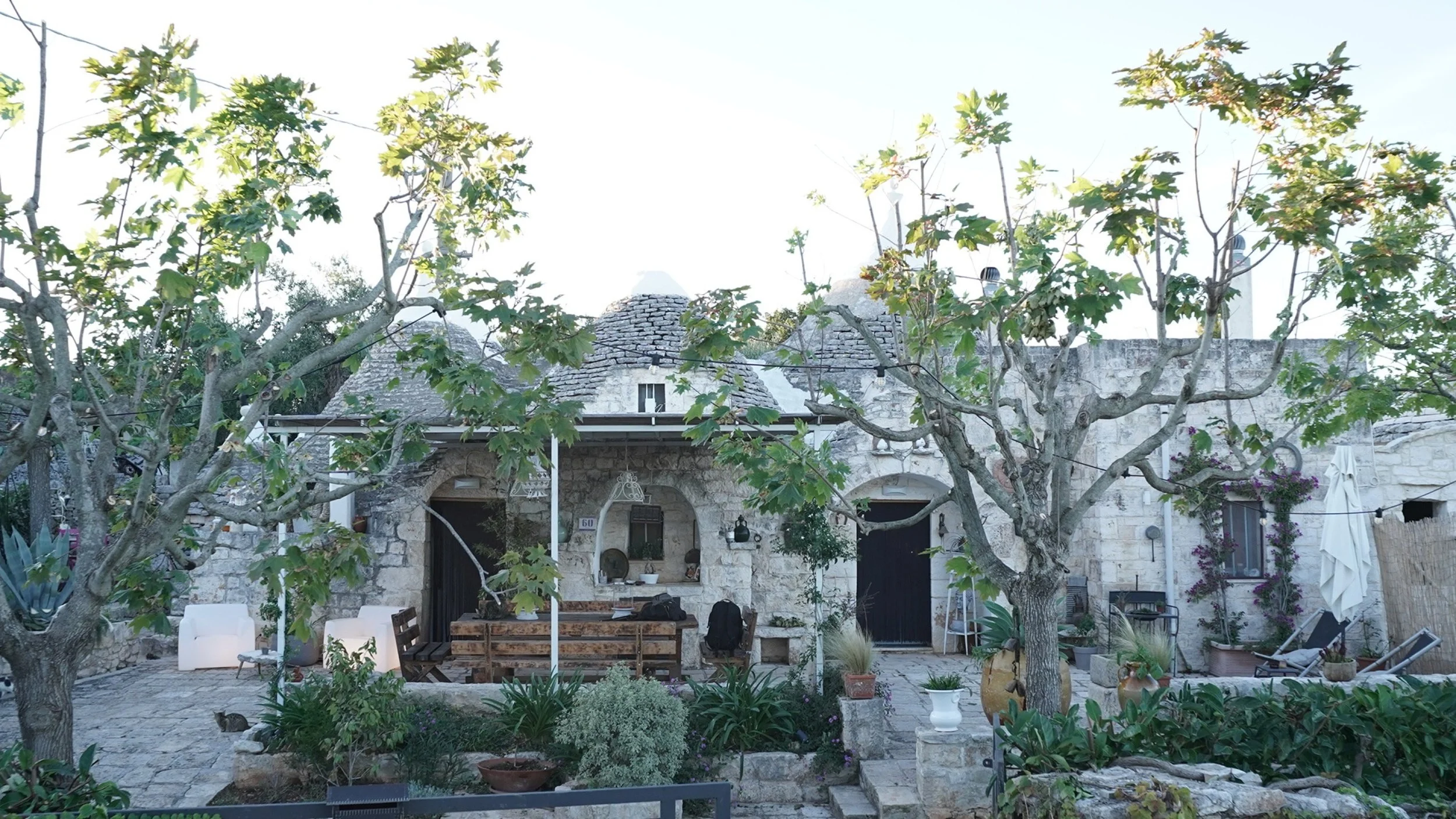 Casa in pietra con tetti conici in pietra, due alberi con foglie verdi, panchine e sedie nel cortile, piante e vasi decorativi, in un ambiente tranquillo e rustico.