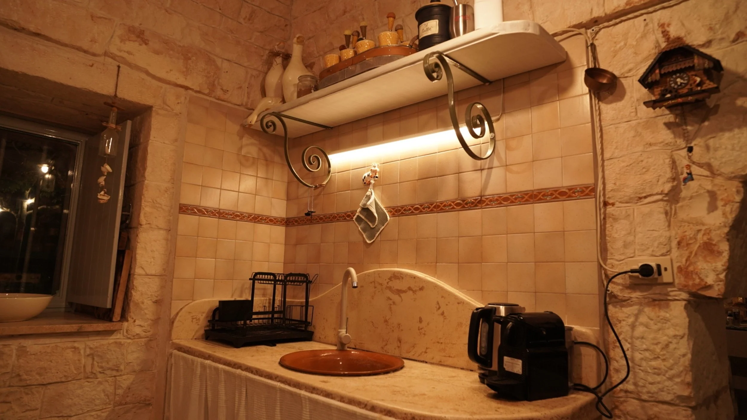 Angolo di una cucina rustica con pareti in pietra e piastrelle, lavabo con piano in marmo, scaffale con bottiglie e barattoli, macchine da caffè e altri piccoli elettrodomestici.