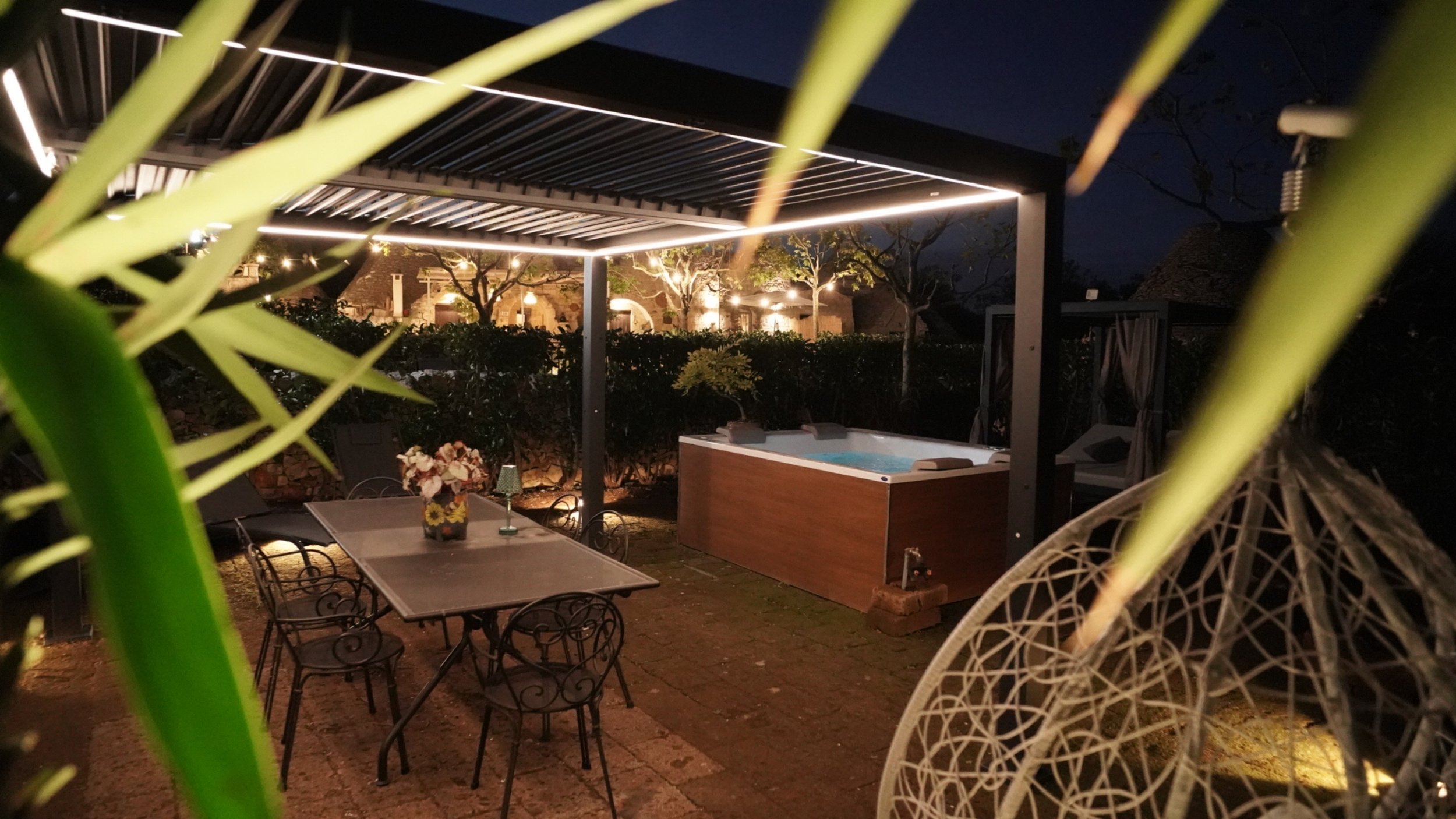 Spazio esterno notturno con tavolo, sedie, jacuzzi, area relax con amaca e illuminazione decorativa.