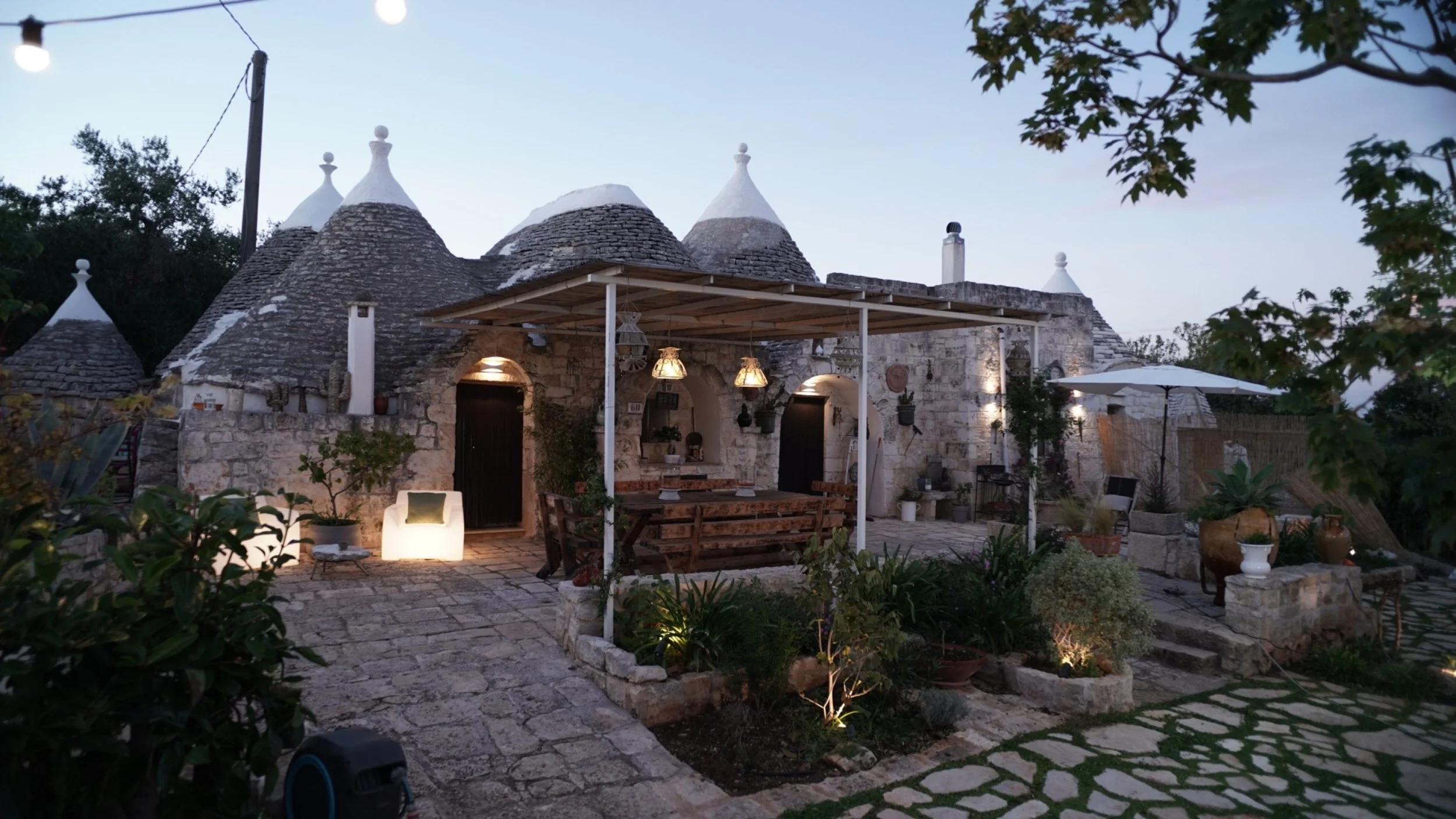 Esterno di una casa in pietra con tetti conici a forma di tronco di cono, illuminazione calda e un'area esterna con arredamento, piante e luci soffuse in serata.