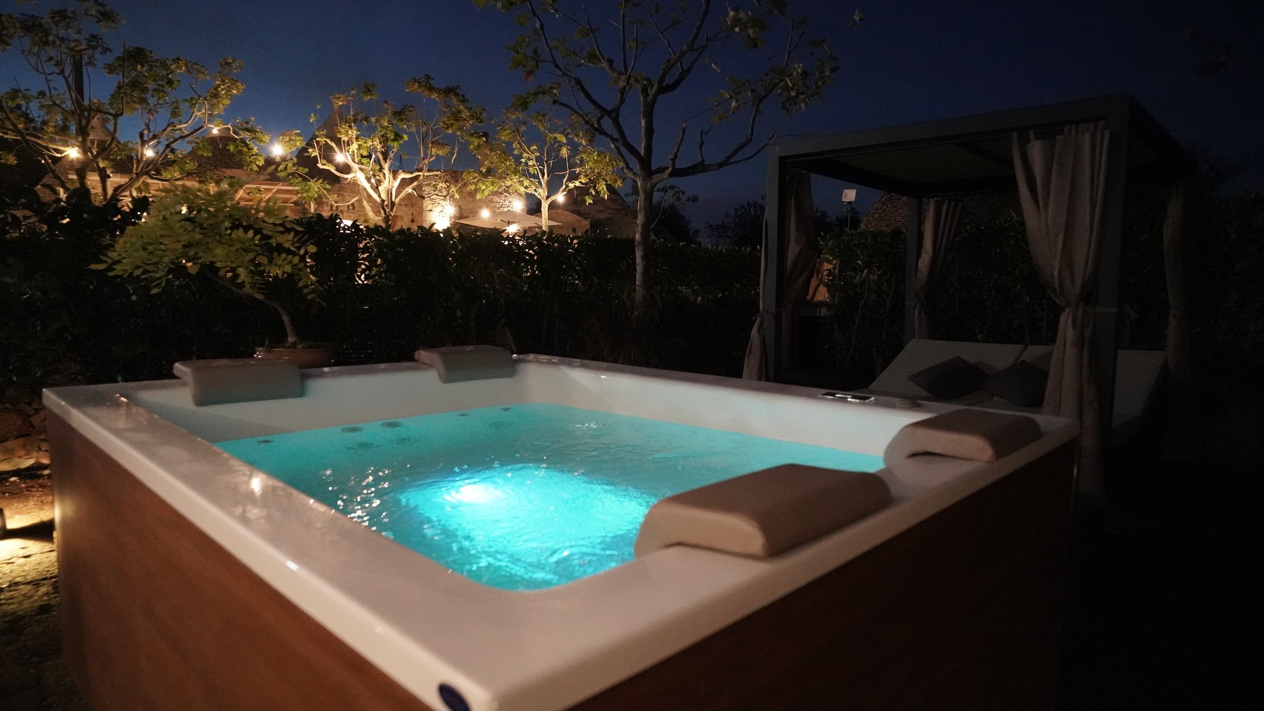 Una spa all'aperto con jacuzzi illuminato di notte, circondato da alberi e una postazione con cuscini e tende.