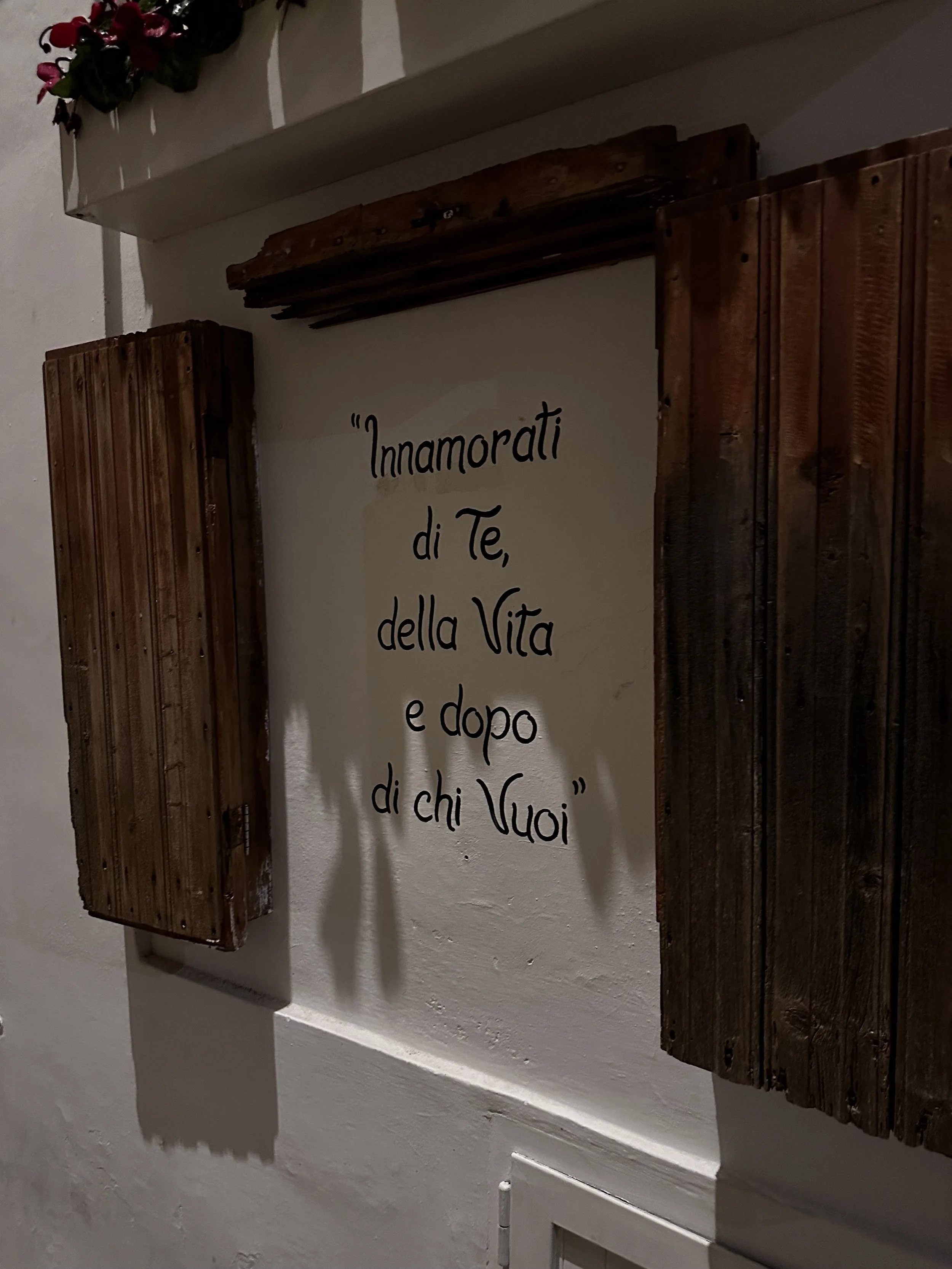 Muro bianco con citazione in italiano: "Innamorati di Te, della Vita e dopo di chi Vuoi" e decorazioni di legno.