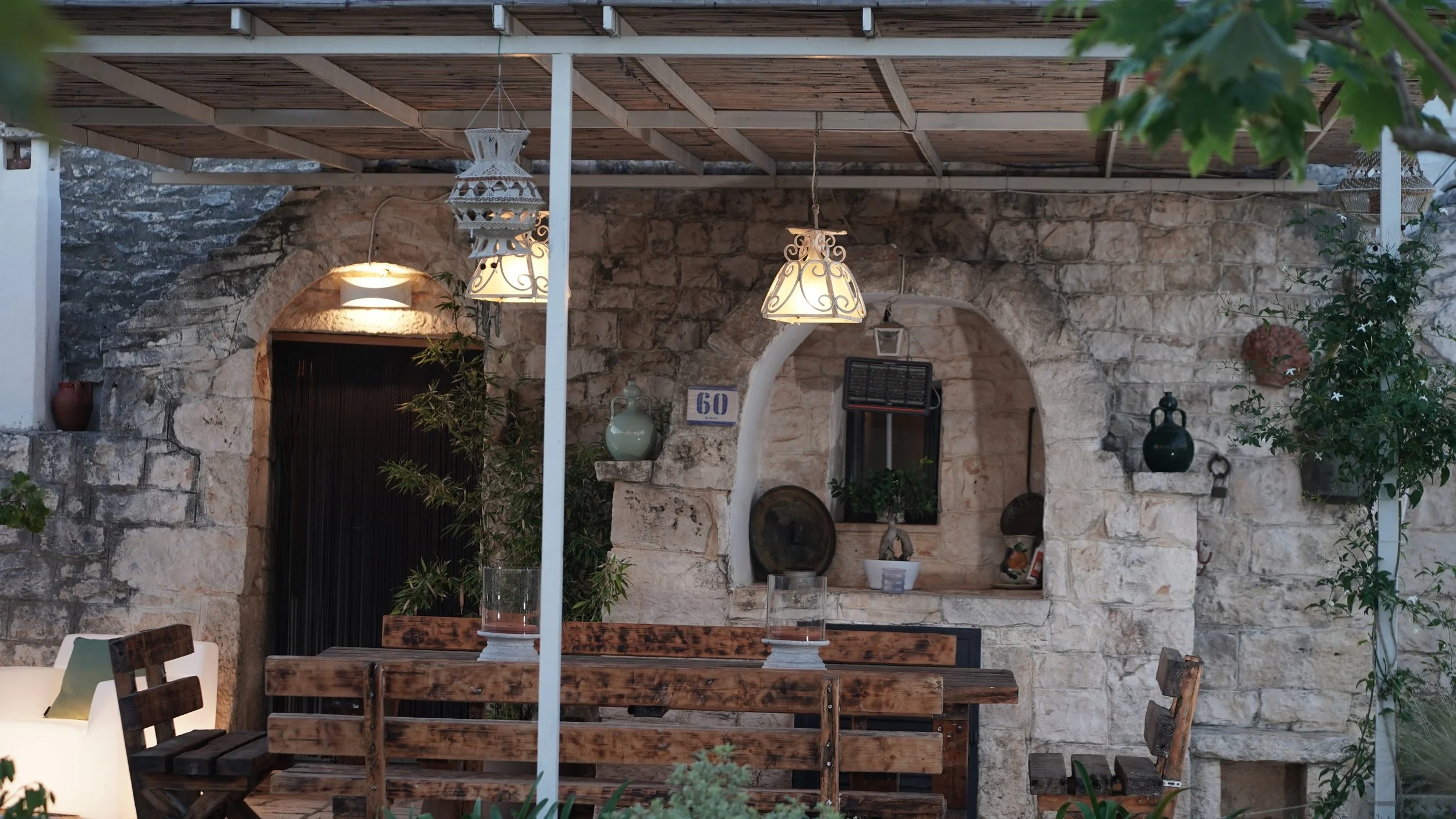 Esterno di una terrazza in pietra con tavolo in legno, sedie e lampade decorative, decorata con piante e vasi, con un arco in pietra e un muro di pietra sullo sfondo.
