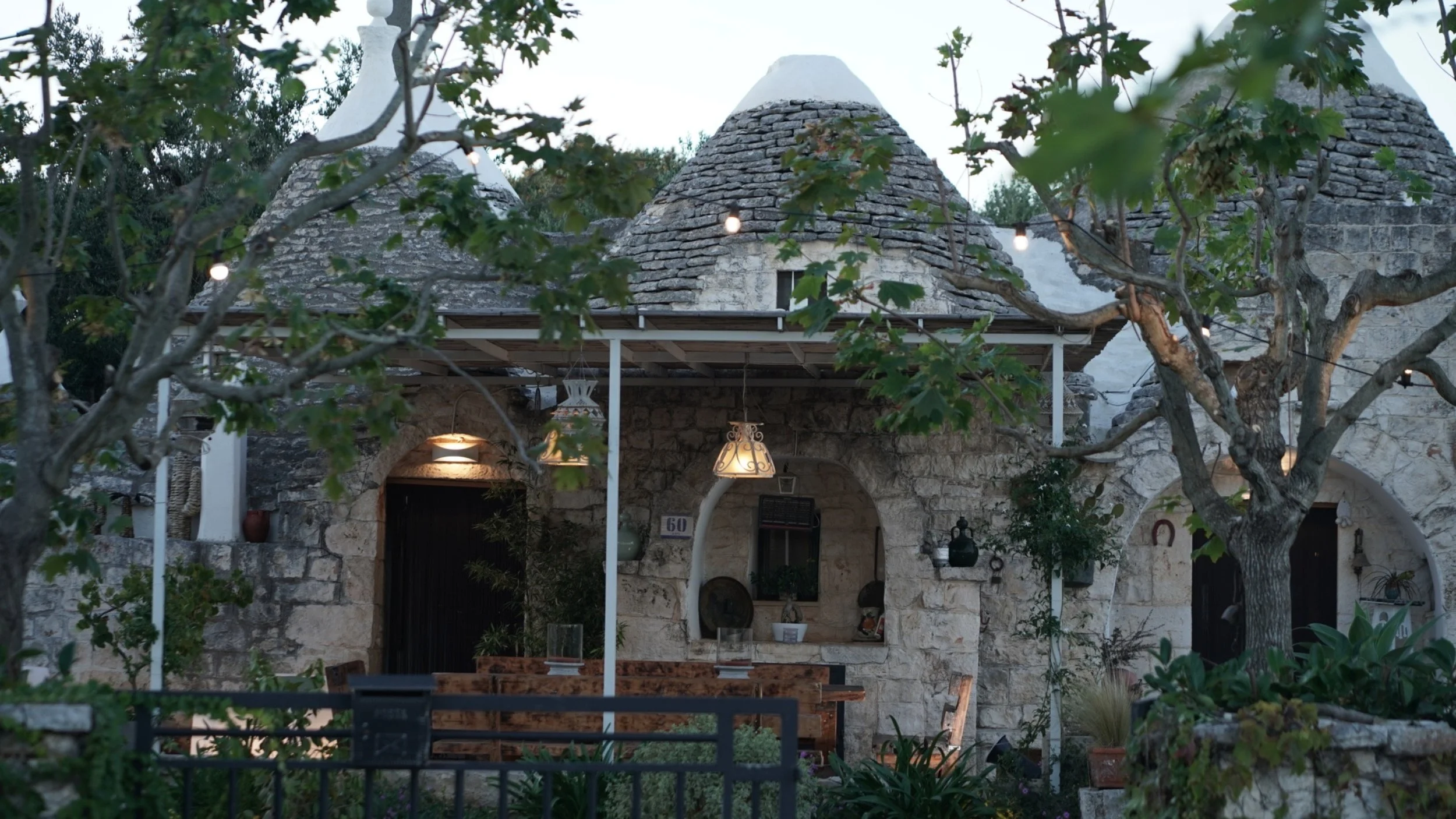 Casa di pietra in stile trullo con tetto conico, circondata da alberi e piante, con luci esterne accese e arredi da giardino.