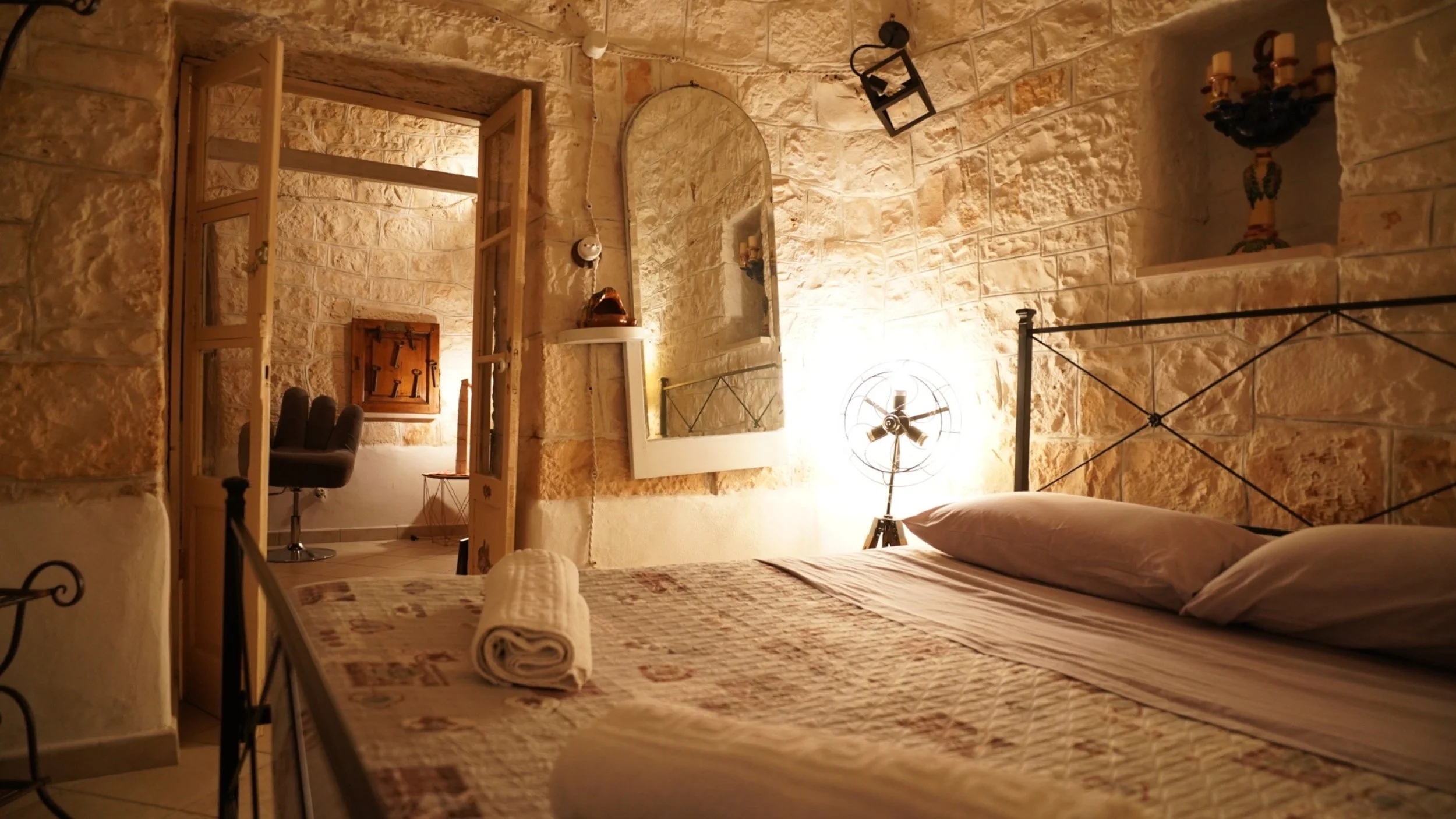 Camera da letto con pareti di pietra, letto con lenzuola e cuscini, specchio sulla parete, ventilatore in piedi e lampada vintage, in una stanza accogliente e luminosa.