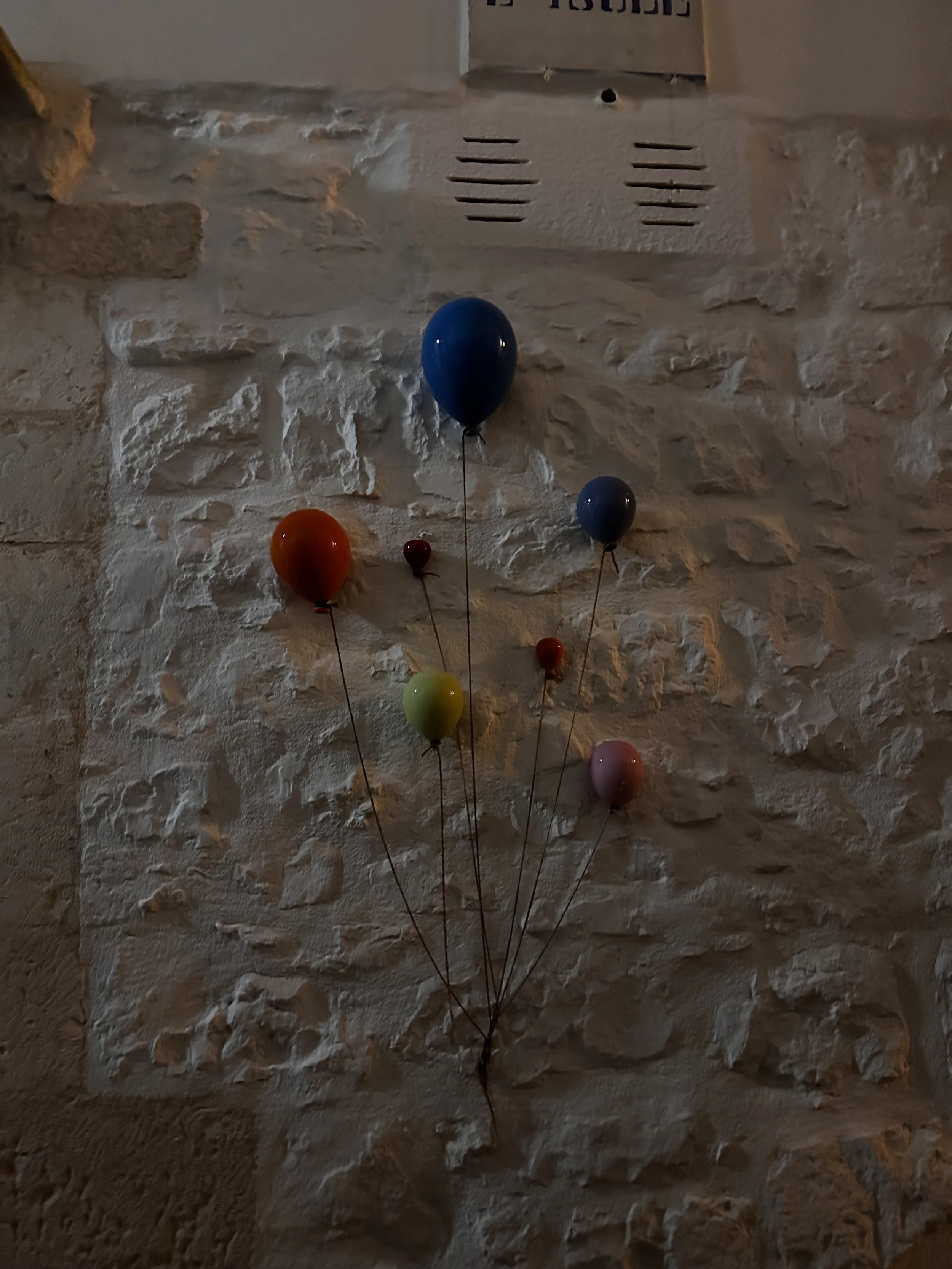 Un muro di pietra con dei palloncini colorati attaccati con dei fili, tra cui palloncini blu, arancione, giallo, rosso, rosa e più piccoli.