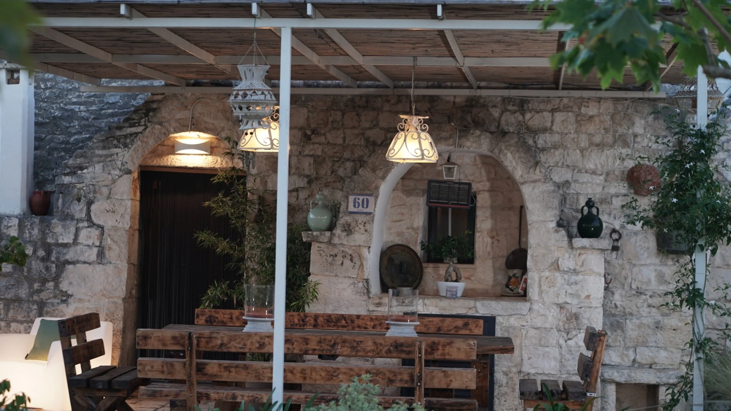 Interno di una casa rustica con pareti di pietra, tavolo in legno con sedie, arredamento antico e lampade decorative sospese piene di luce calda, piante e vasi decorativi.