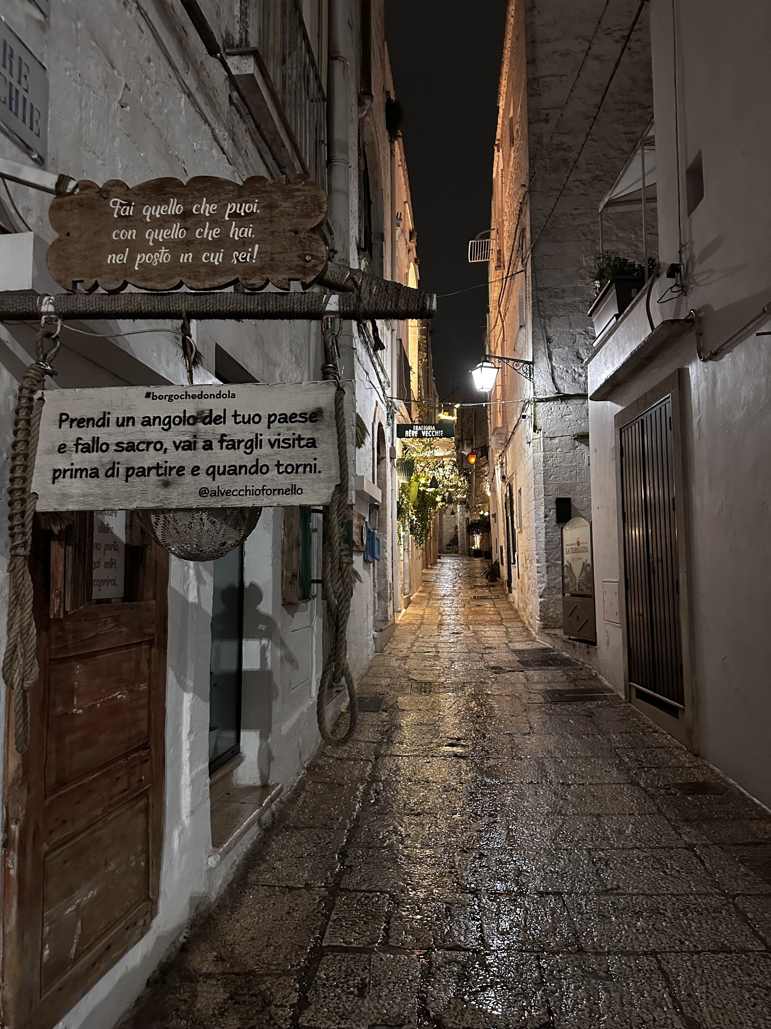 Una via stretta con edifici in pietra e illuminazione notturna, con segnali di scritte in italiano e luci lungo la strada.