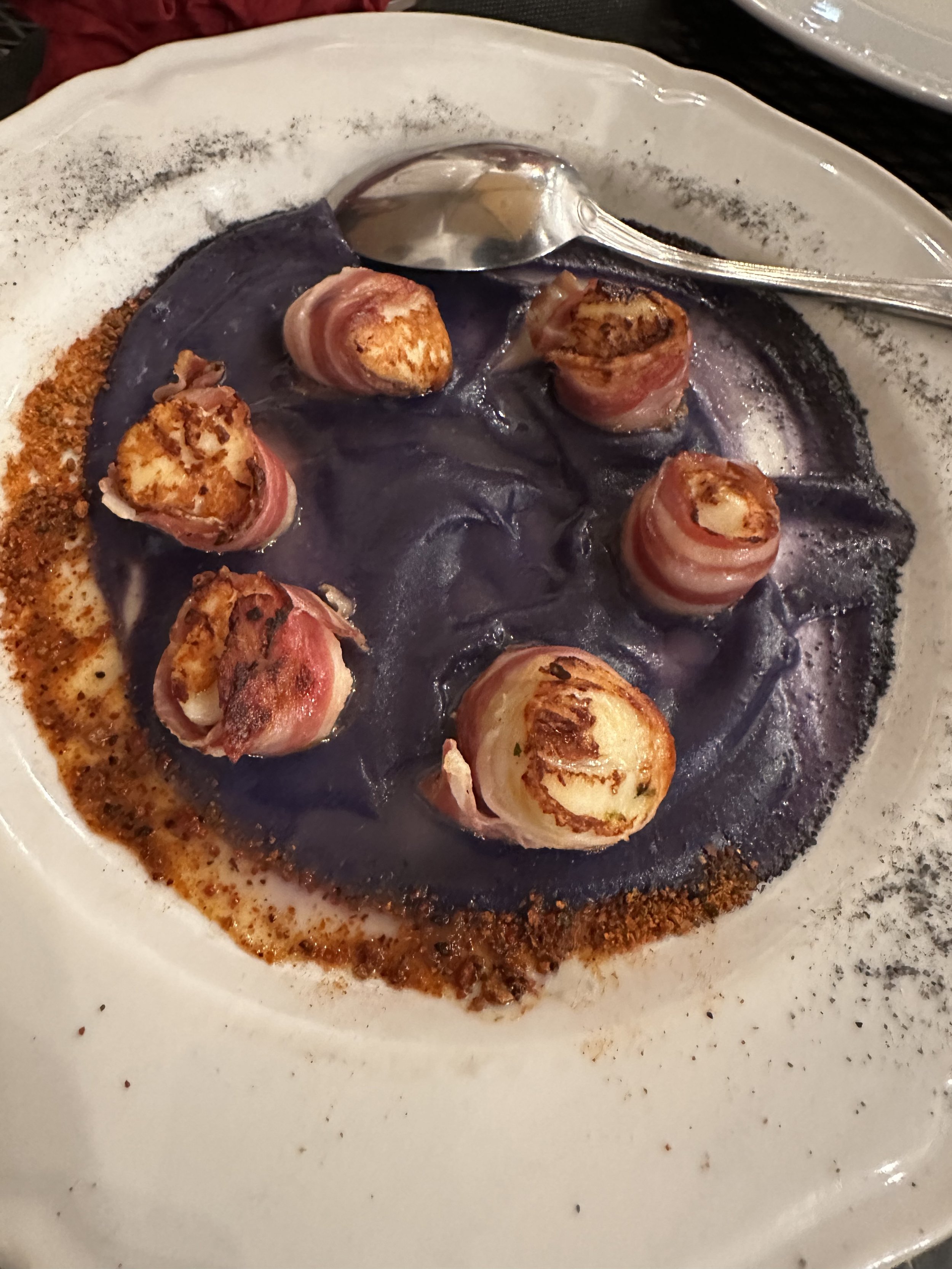 Piatti di cozze ripiene avvolte in pancetta, servite con purea di patate viola e una spezia in polvere sul bordo del piatto.