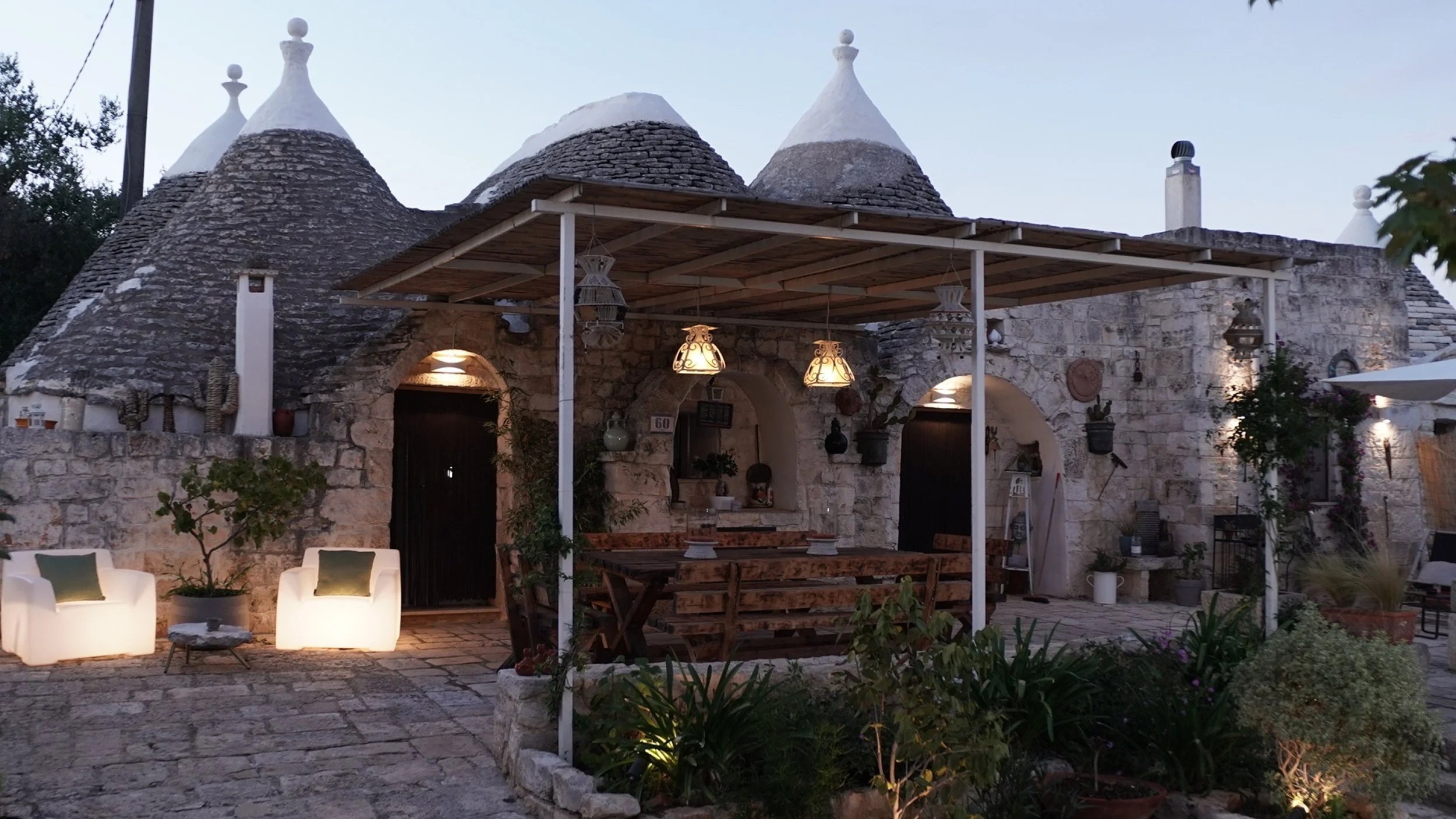 Casa in pietra con tetti conici tipici dei trulli, illuminata di sera con arredi esterni e piante decorate, atmosfera accogliente.