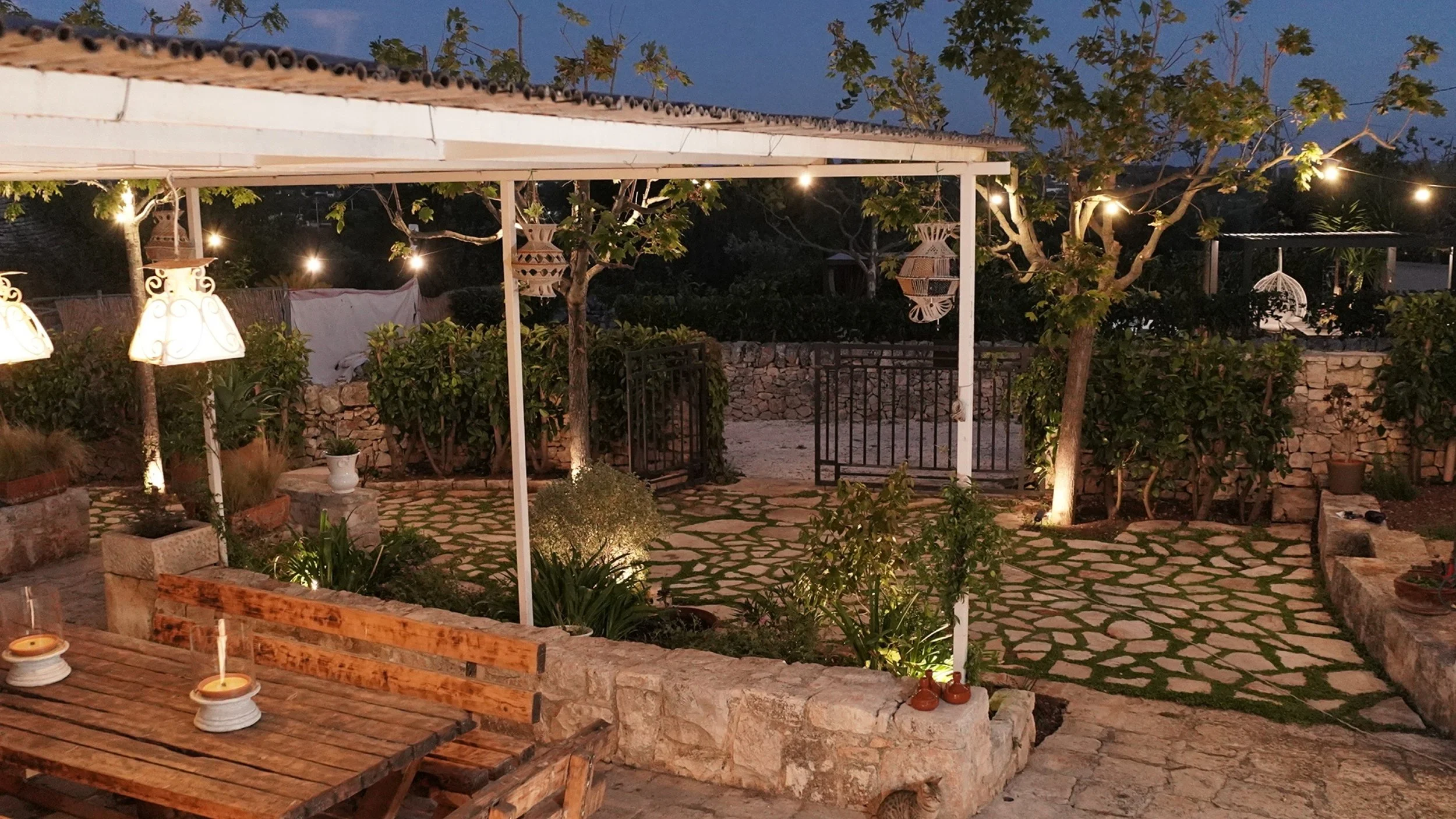 Giardino esterno decorato con luci soffuse, piante, banci in legno, lampade e gabbie decorative, con un cancello nero in metallo e un muro di pietra sullo sfondo.