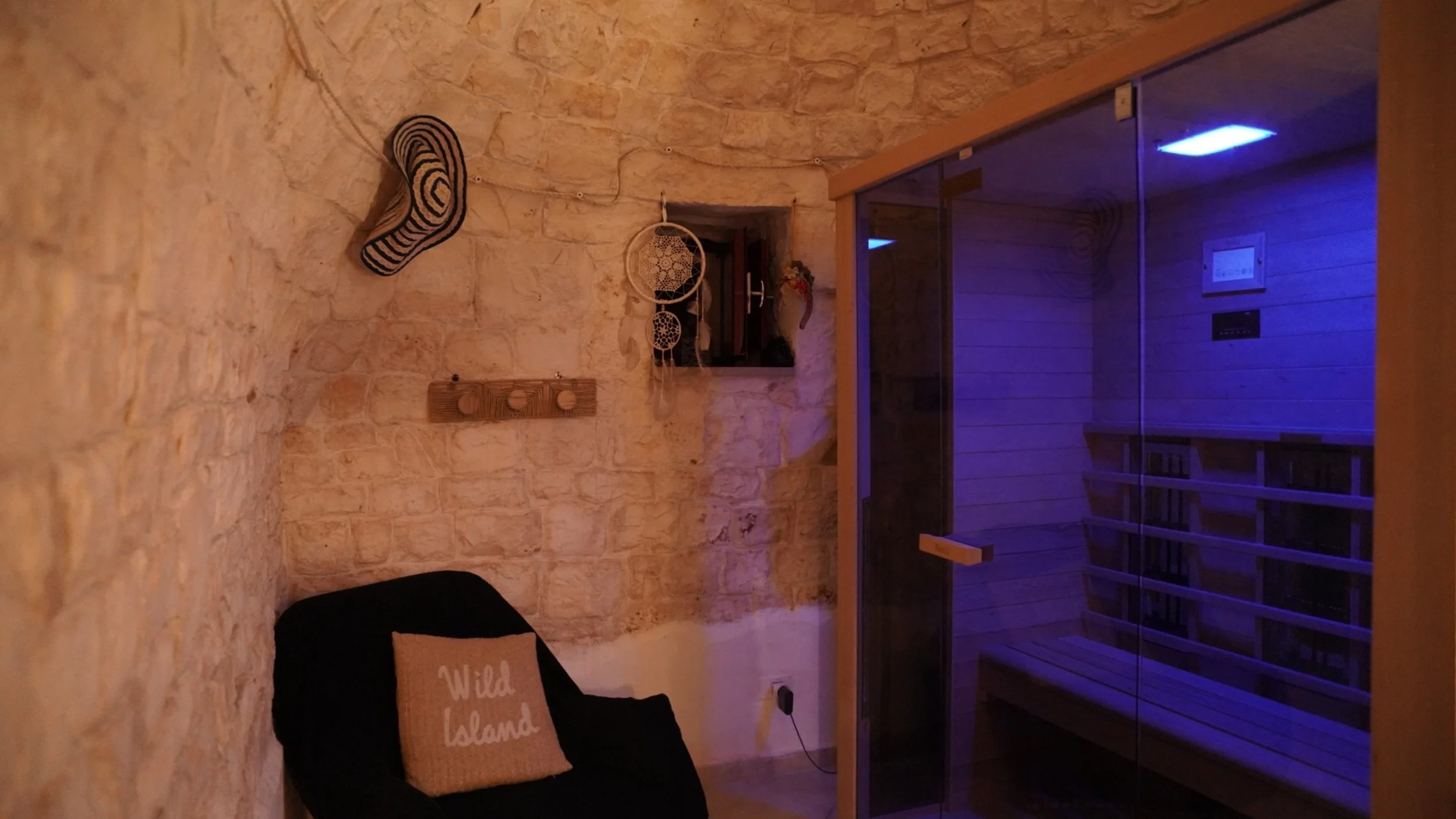 Stanza con pareti di pietra, poltrona nera con cuscino 'Wild Island', parete con decorazioni di soggetti in velluto, sauna in vetro con illuminazione blu, pareti di legno, scaffale con libri