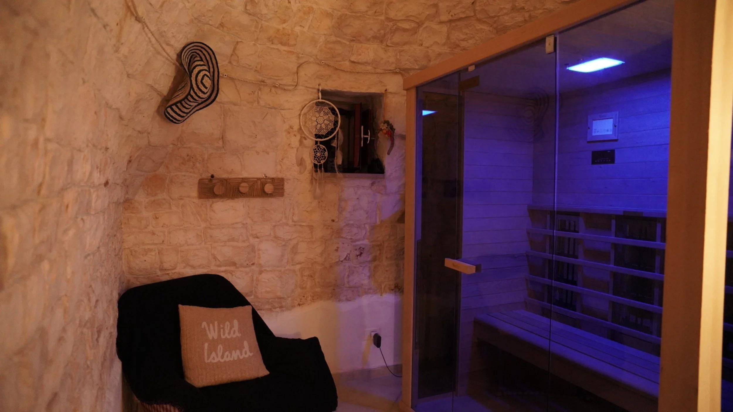 Stanza con pareti in pietra, un'insegna a forma di piede e un soggetto appeso con un motivo a nido d'ape, una finestra aperta. Arredamento minimalista con una poltrona nera e un cuscino con scritta 'Wild Island'. Sauna in vetro con luce blu all'inter