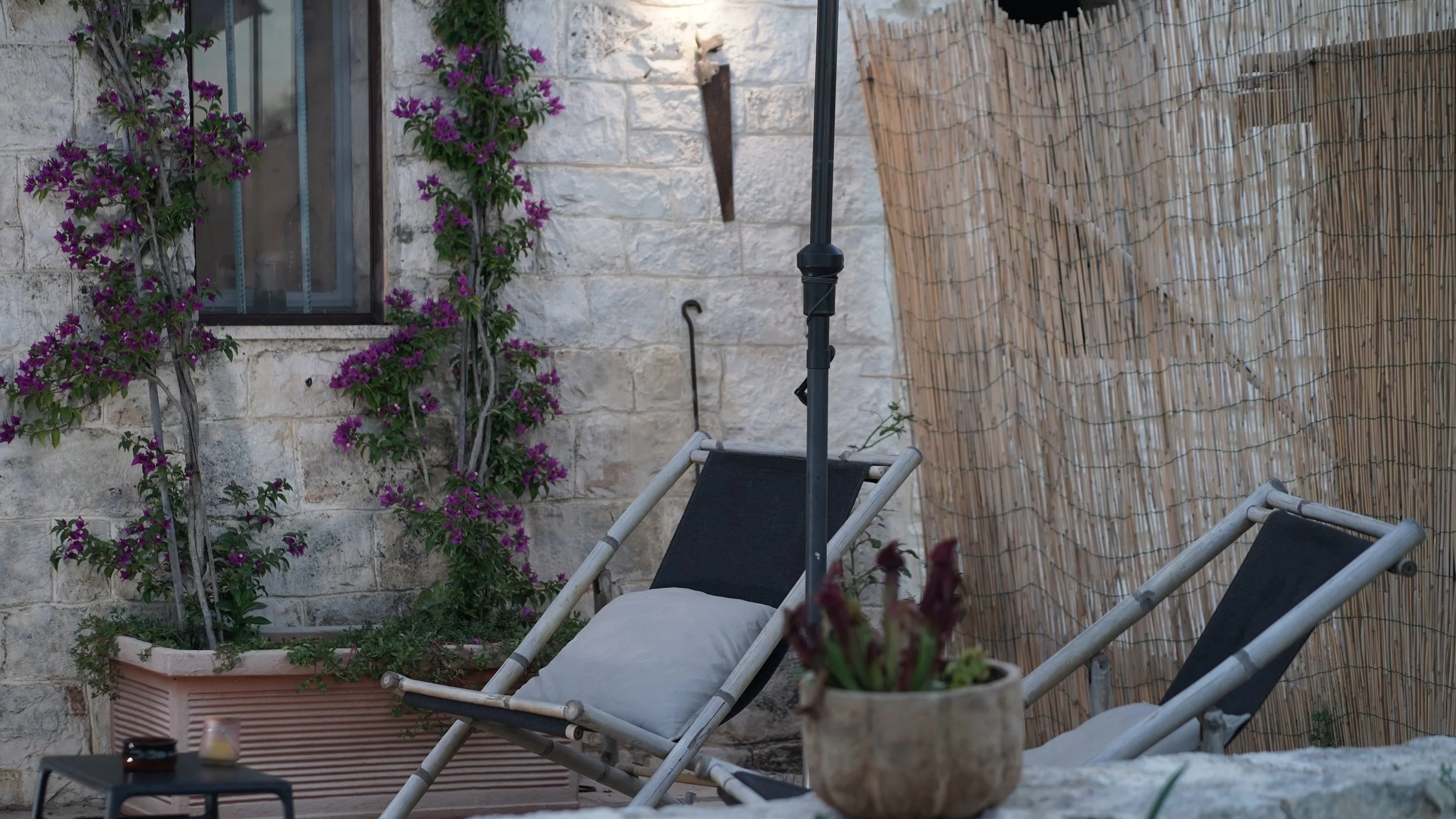 Zona relax all'aperto con due chaise longue nere, un ombrellone chiuso, piante fiorite in vaso e un muro di pietra con una finestra. Texture di recinzione in paglia di fronte.