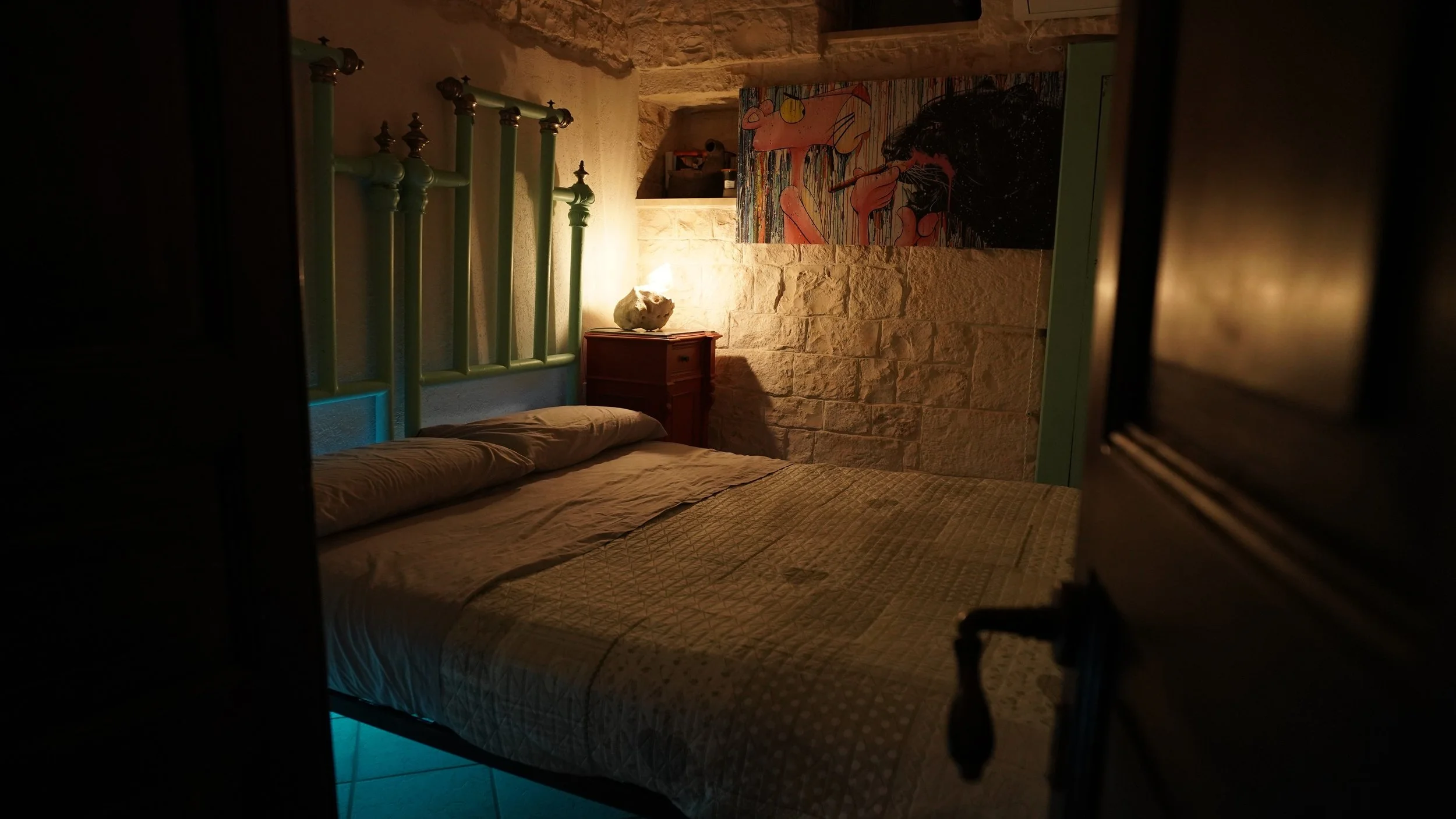 Camera da letto con pareti in pietra, letto con coperte beige, lampada accesa su una cassettiera di legno, dipinto astratto sulla parete