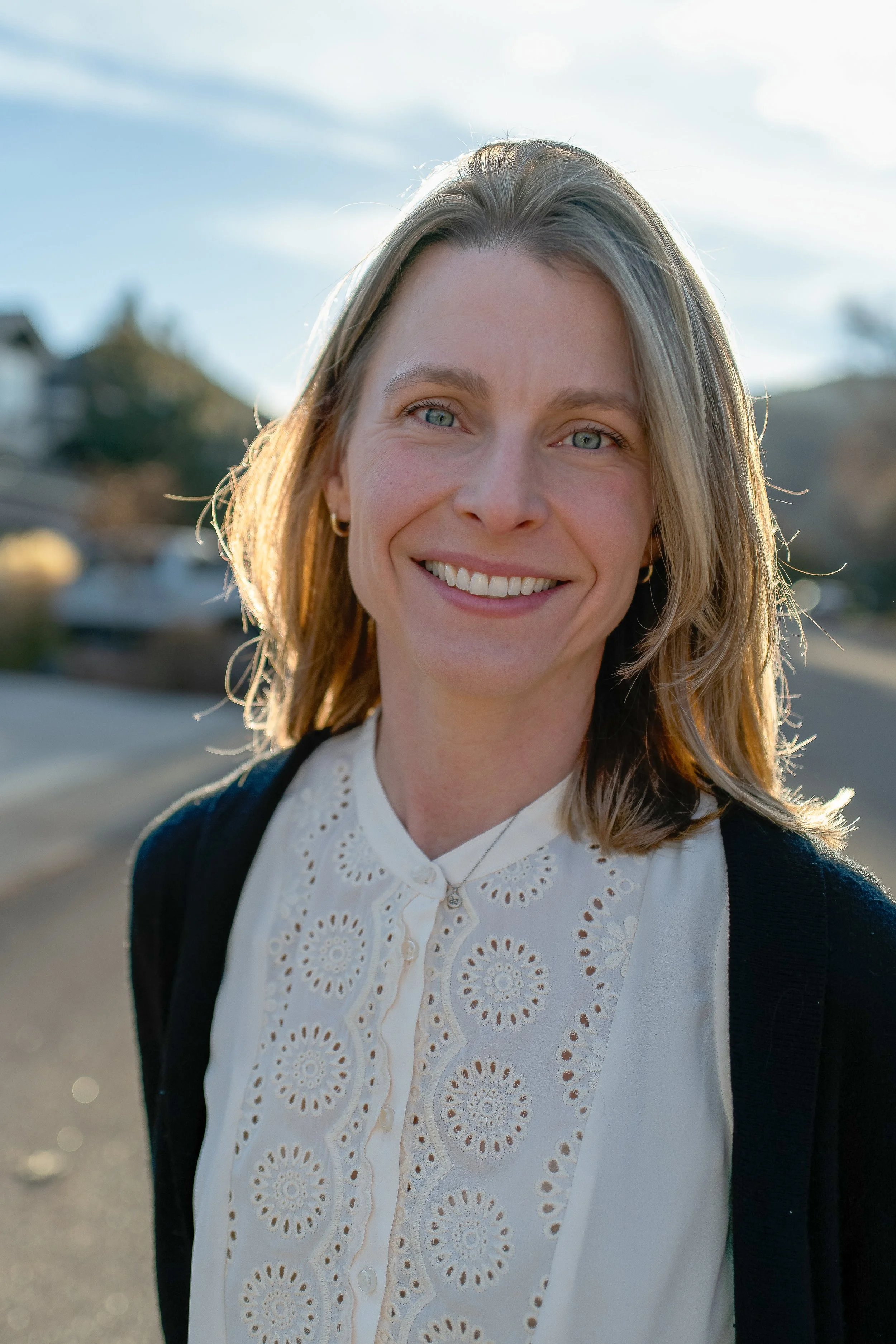 A smiling woman with shoulder-length blonde hair, wearing a white embroidered blouse and a black cardigan, standing outdoors on a sunny day.