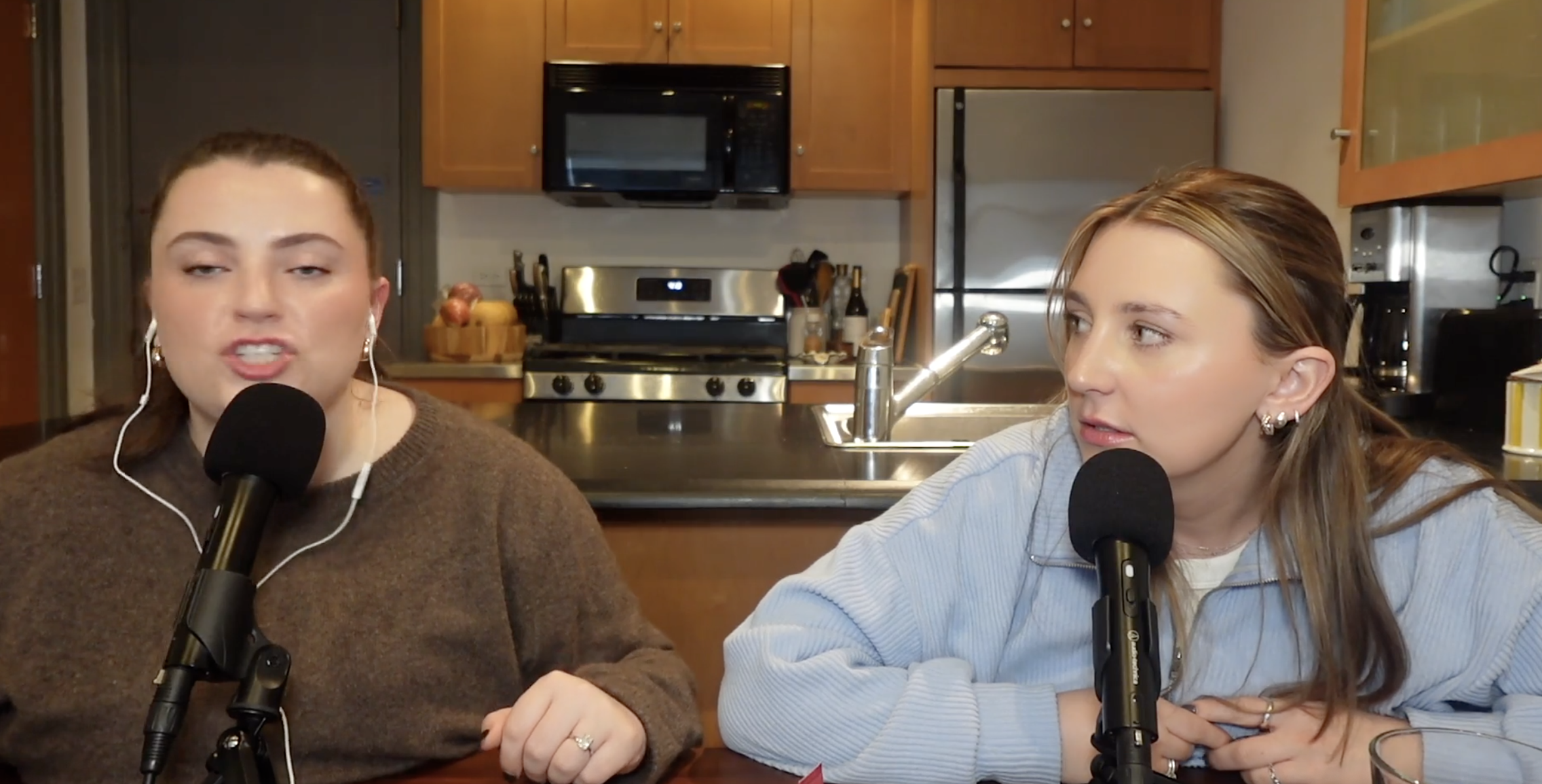 Two women speaking into microphones during a podcast recording in a kitchen setting.
