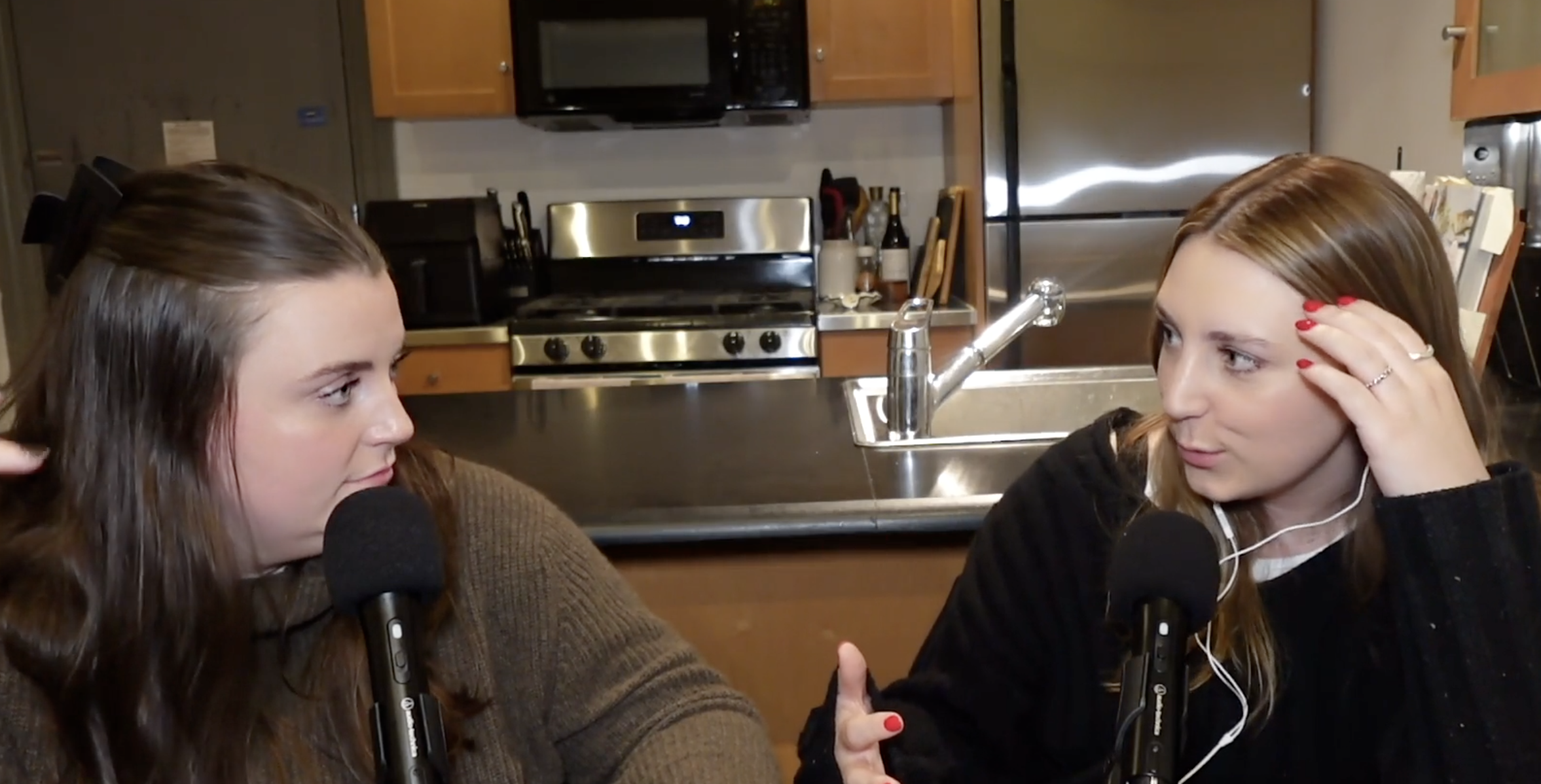Two women are sitting at a kitchen counter, speaking into microphones with headphones on. The woman on the left has long brown hair, and the woman on the right has shoulder-length blond hair, talking and touching her hair. The kitchen in the background has wooden cabinets, a stove, a microwave, and a refrigerator.