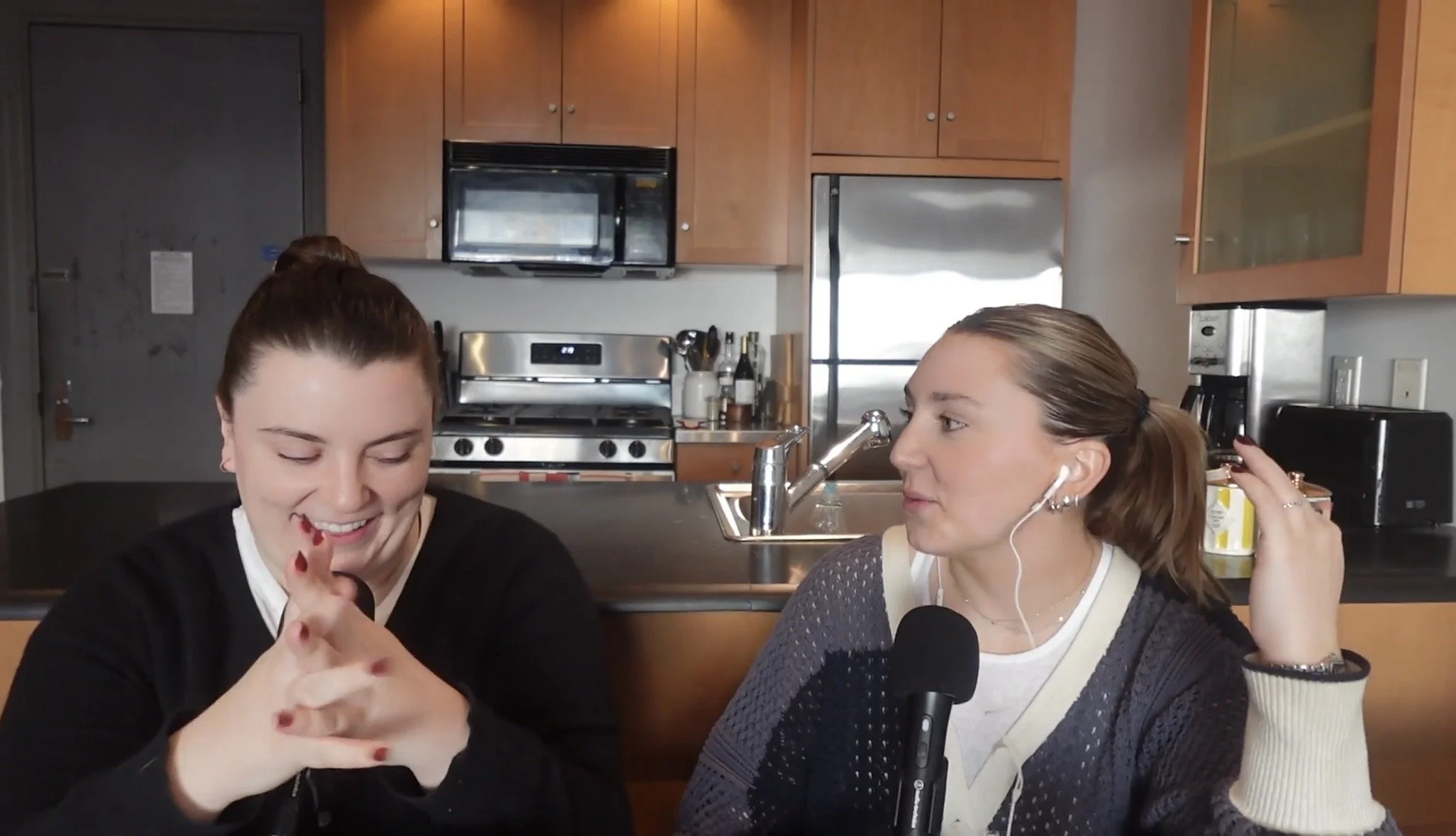 Two women sitting in a kitchen during a podcast or interview, with one smiling and looking down, and the other speaking into a microphone with earphones, kitchen appliances visible in the background.