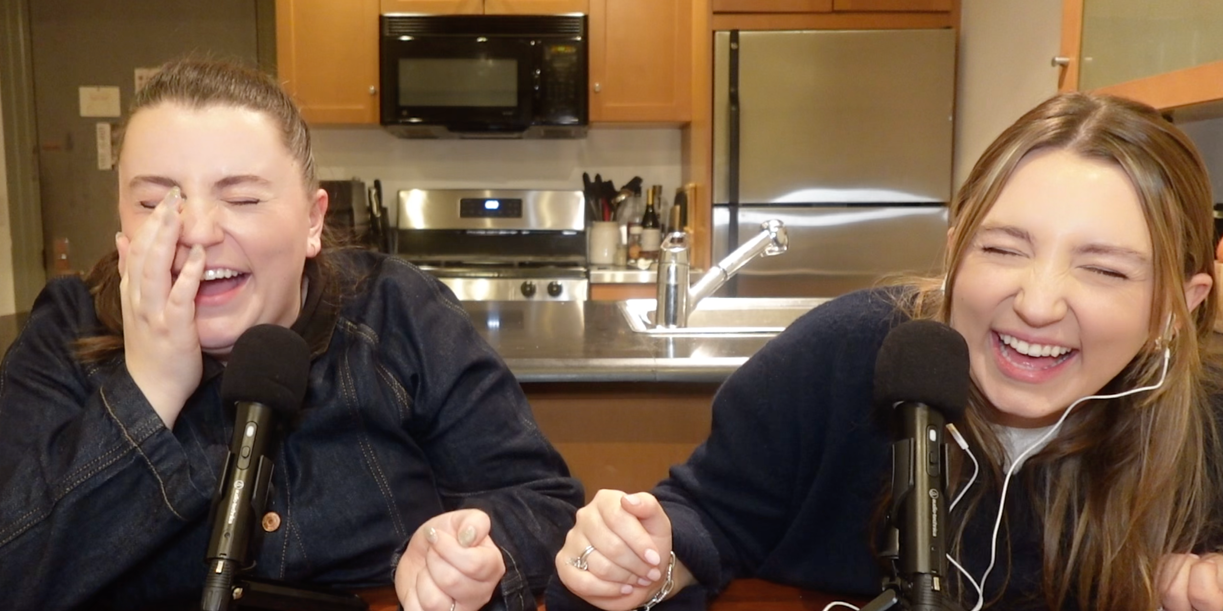 Two women laughing and holding hands in a kitchen with microphones in front of them.