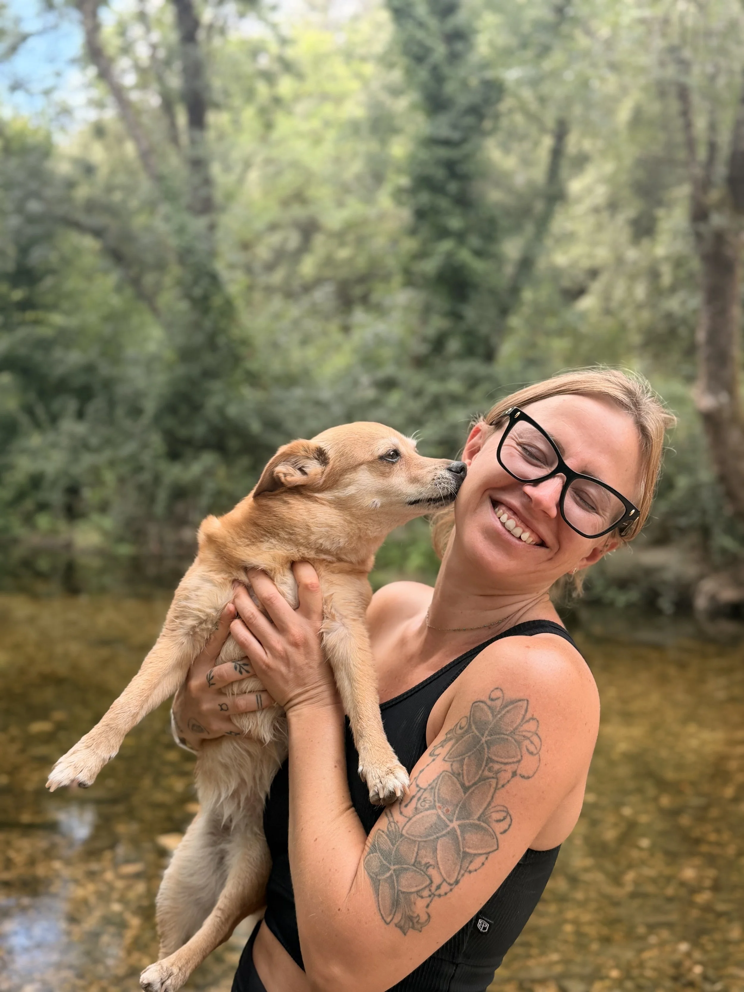 Femme souriante portant un débardeur noir et des lunettes, tenant un chiot brun clair près d'une rivière dans une forêt.