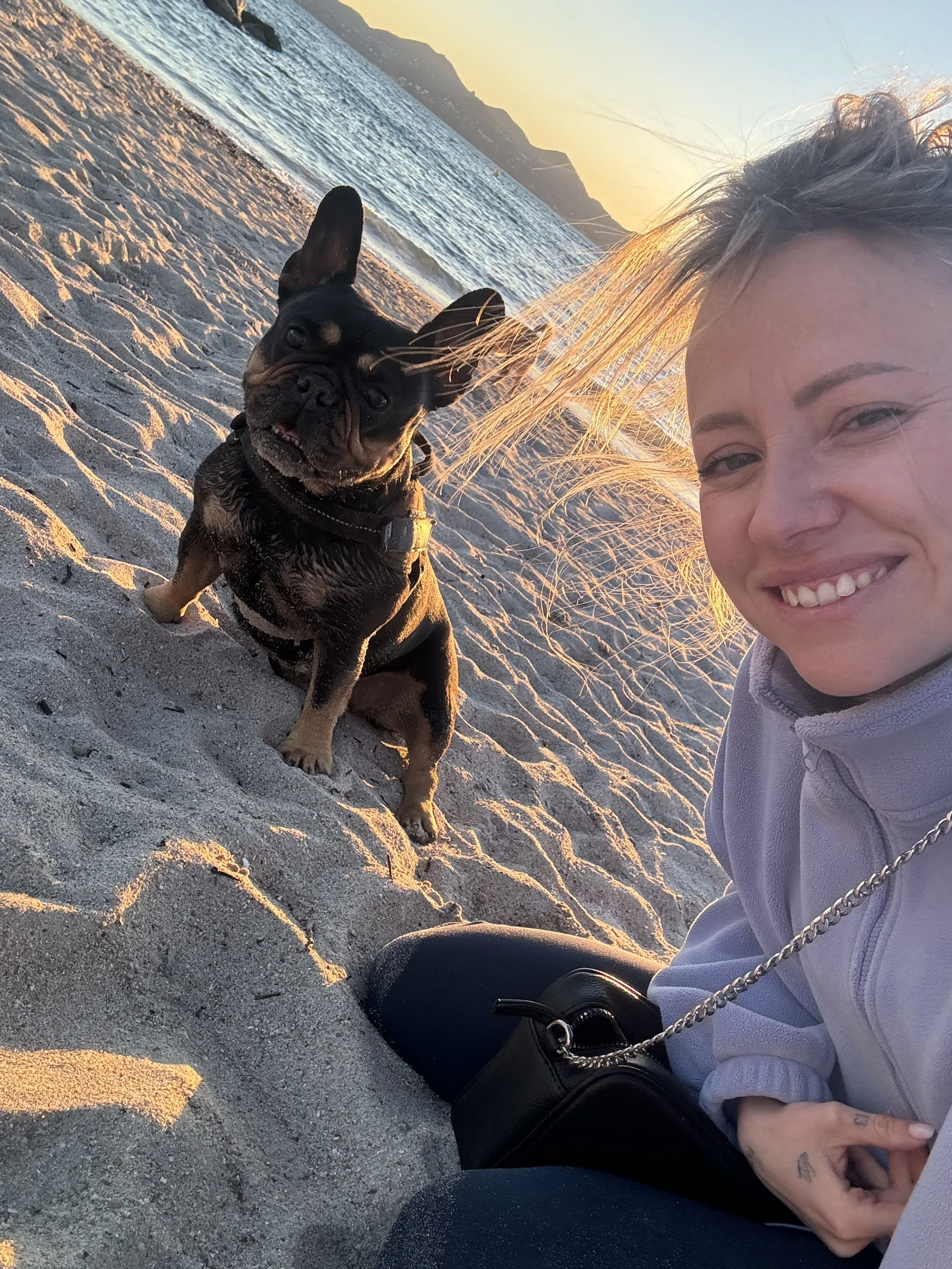 Une femme souriante et son chien French Bulldog assis sur la plage au coucher du soleil.