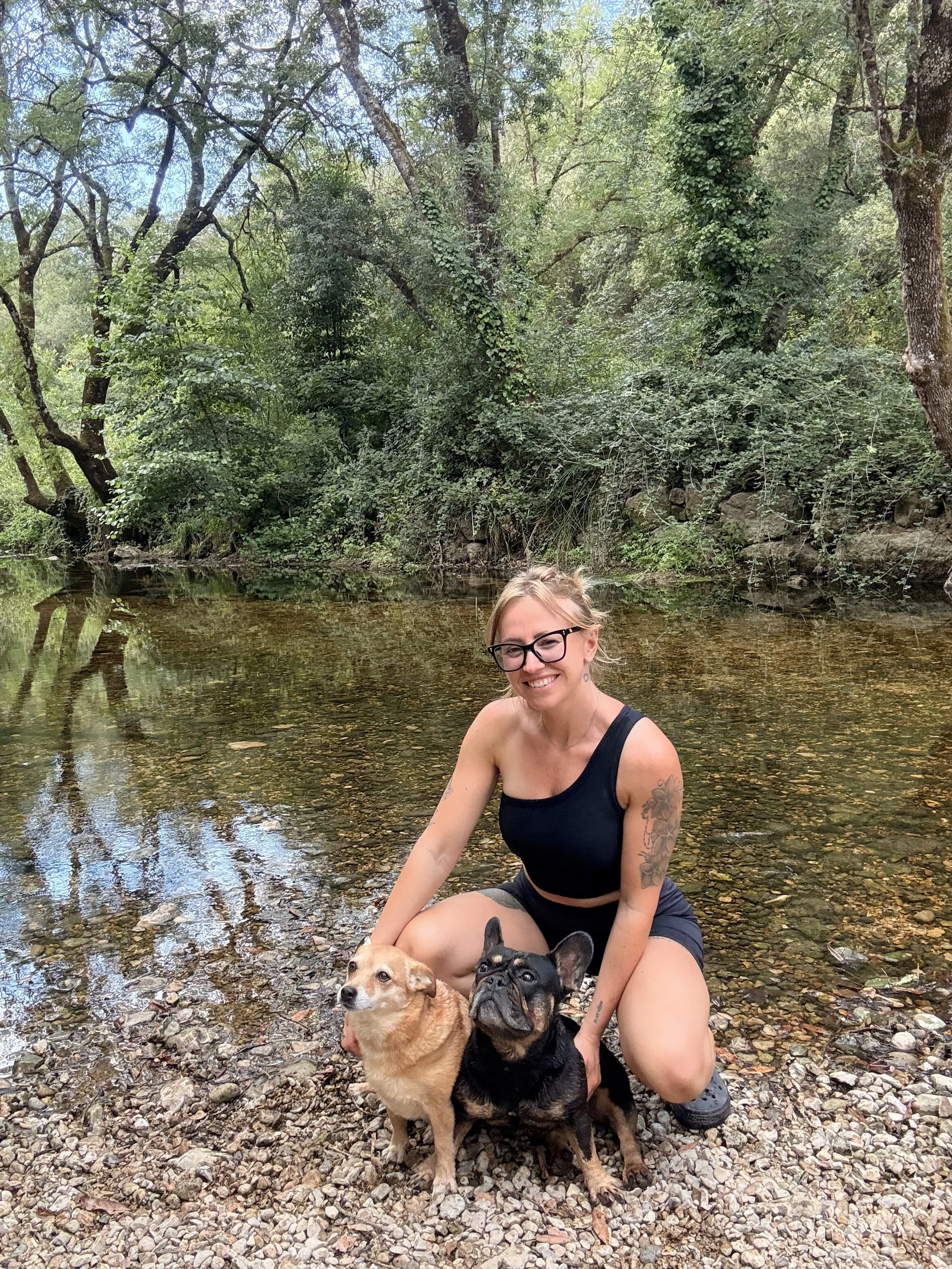 Une femme souriante portant des lunettes noires, en t-shirt noir et shorts, posant avec deux chiens près d'une rivière dans une forêt verdoyante.