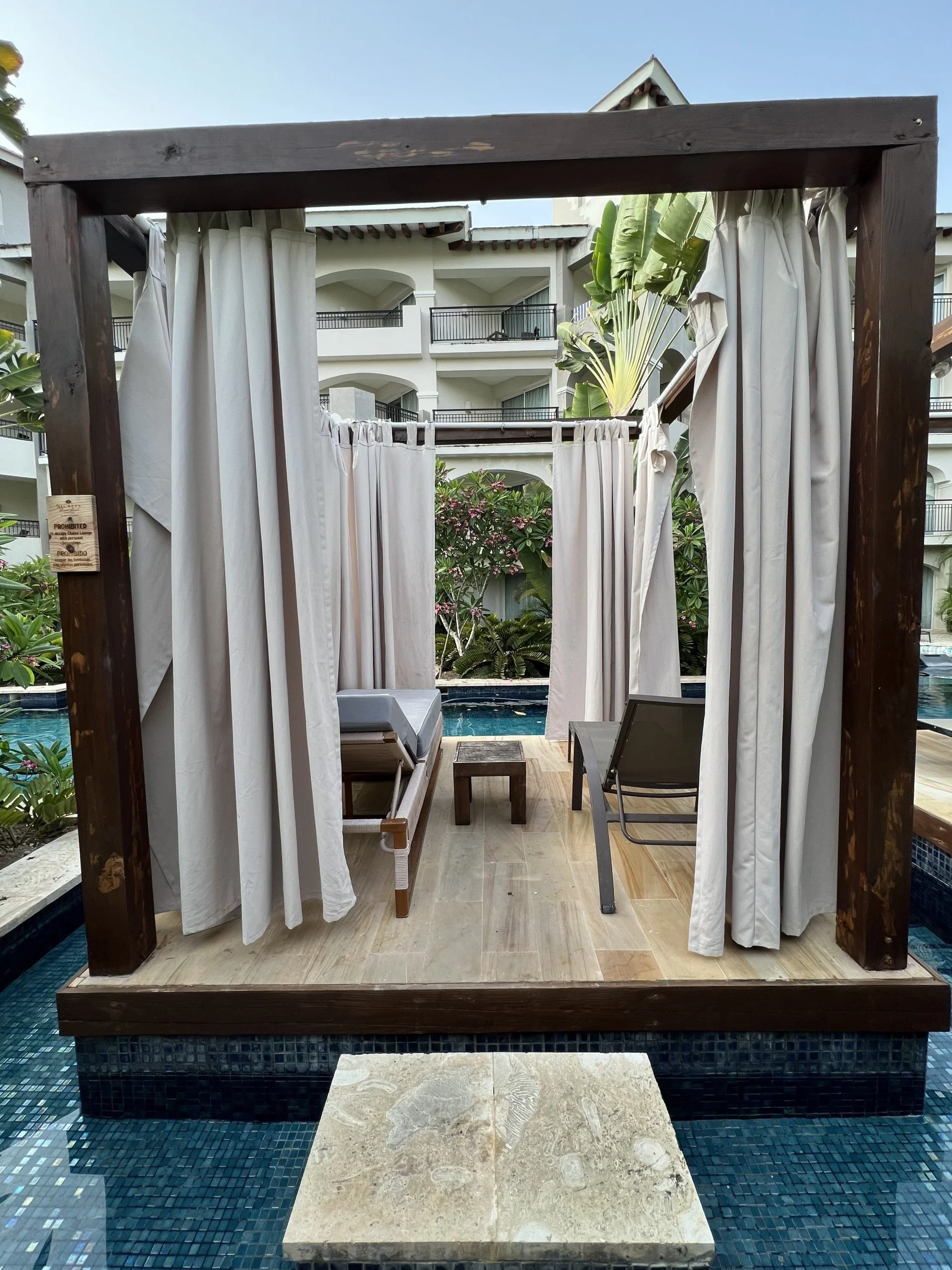 A poolside cabana with white curtains, wooden frame, seating furniture, and a view of a building with balconies and tropical plants in the background.