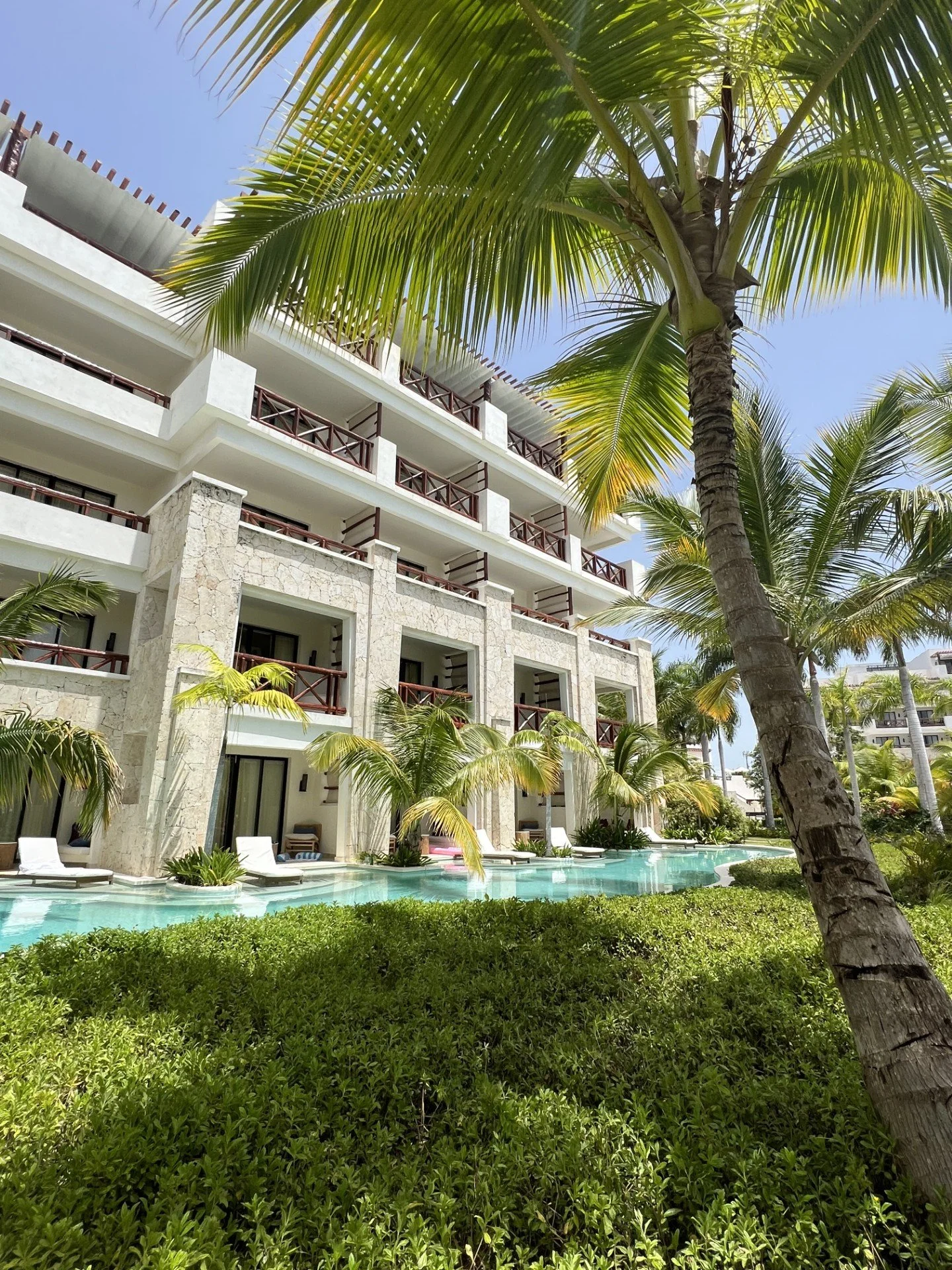 View of a tropical resort with a multi-story white building, balconies with wooden railings, numerous palm trees, Luxury all-inclusive resort vacations with Karen at Wande bushes, and an outdoor pool area with lounge chairs under a partly cloudy sky.