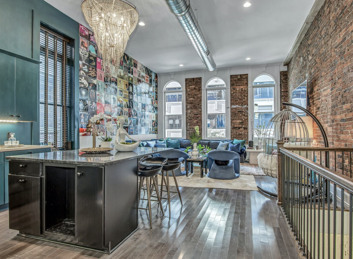 Open-concept living room and kitchen with exposed brick walls, large arched windows, a blue sectional sofa, contemporary chairs, and a gallery wall of colorful artwork.
