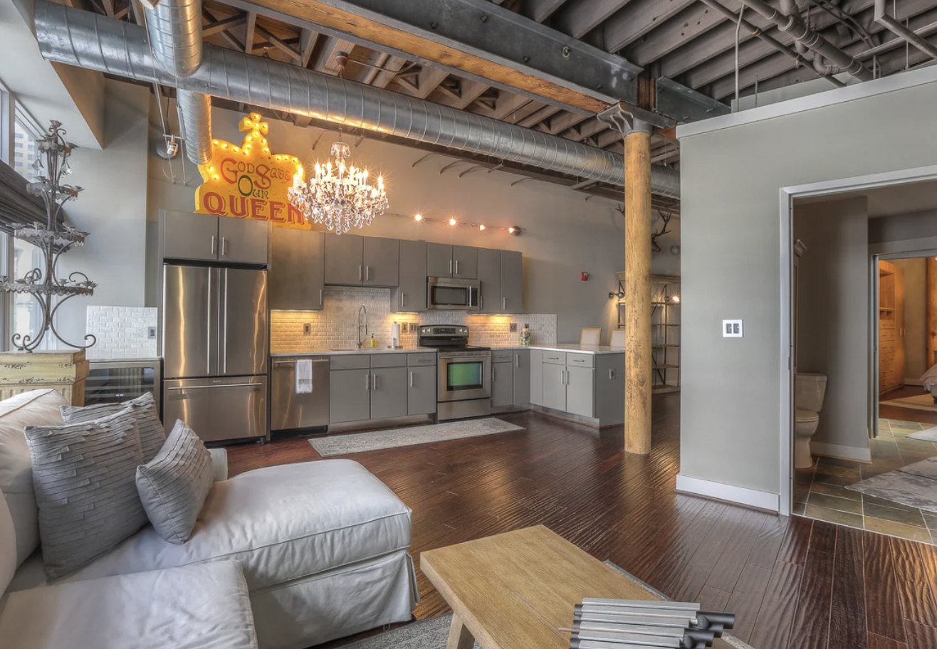 Open-concept kitchen and living area with modern gray cabinets, stainless steel appliances, a chandelier, and a decorative sign that says "God Save Our Queen". Exposed ceiling beams and ductwork are visible, with hardwood floors and a mix of furniture.