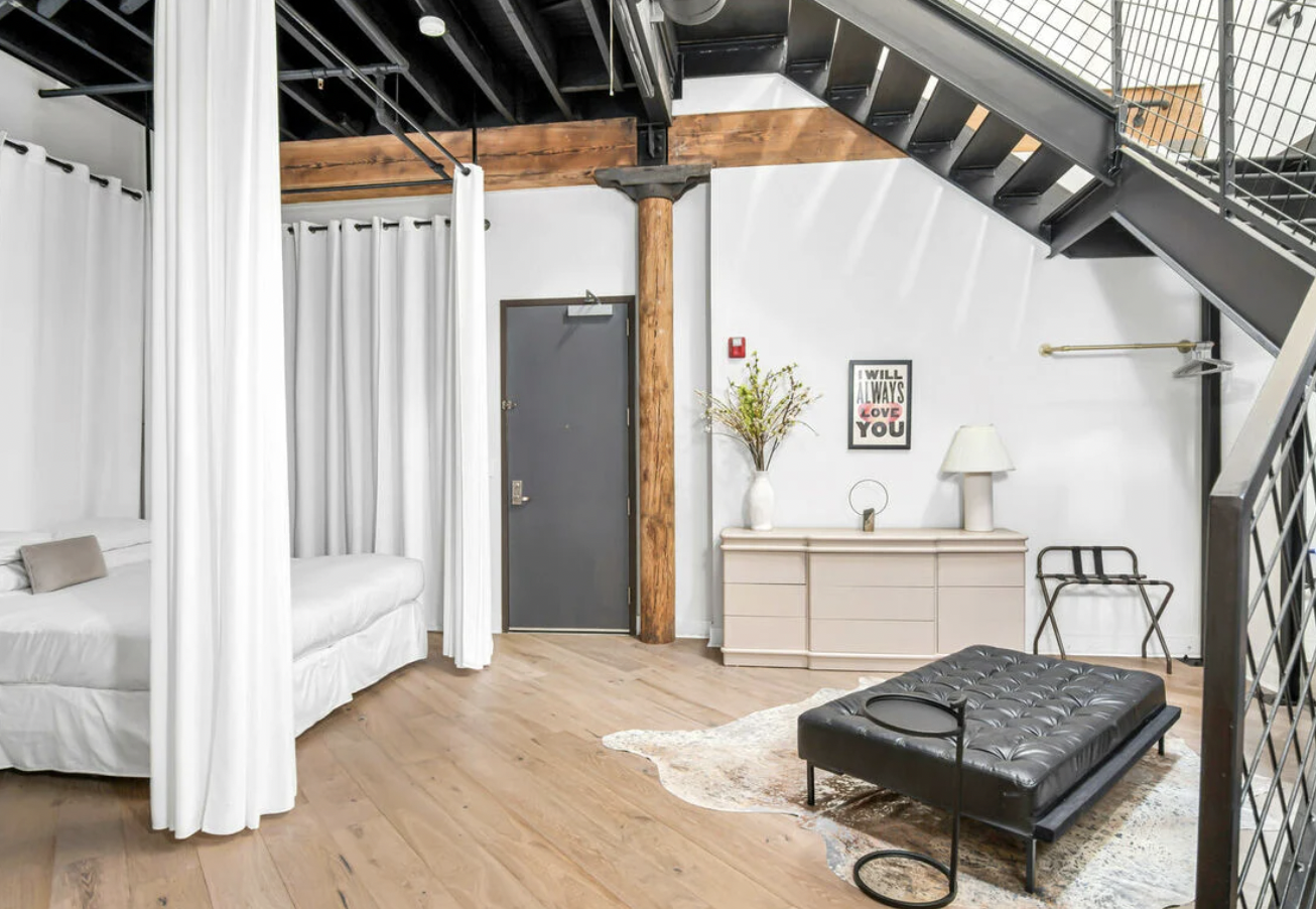 Contemporary living room with a white curtained canopy bed on the left, dark wooden beams and trestle, white walls, and a staircase on the right. A black tufted ottoman is on a patterned rug on a hardwood floor, with a pink dresser topped with a white lamp, a tall vase with flowers, and a framed quote on the wall behind.