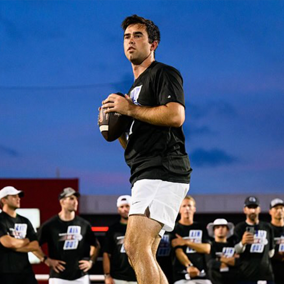 A young man in black sportswear holding a football on a field during dusk, with a group of people in black shirts and white caps watching behind him.