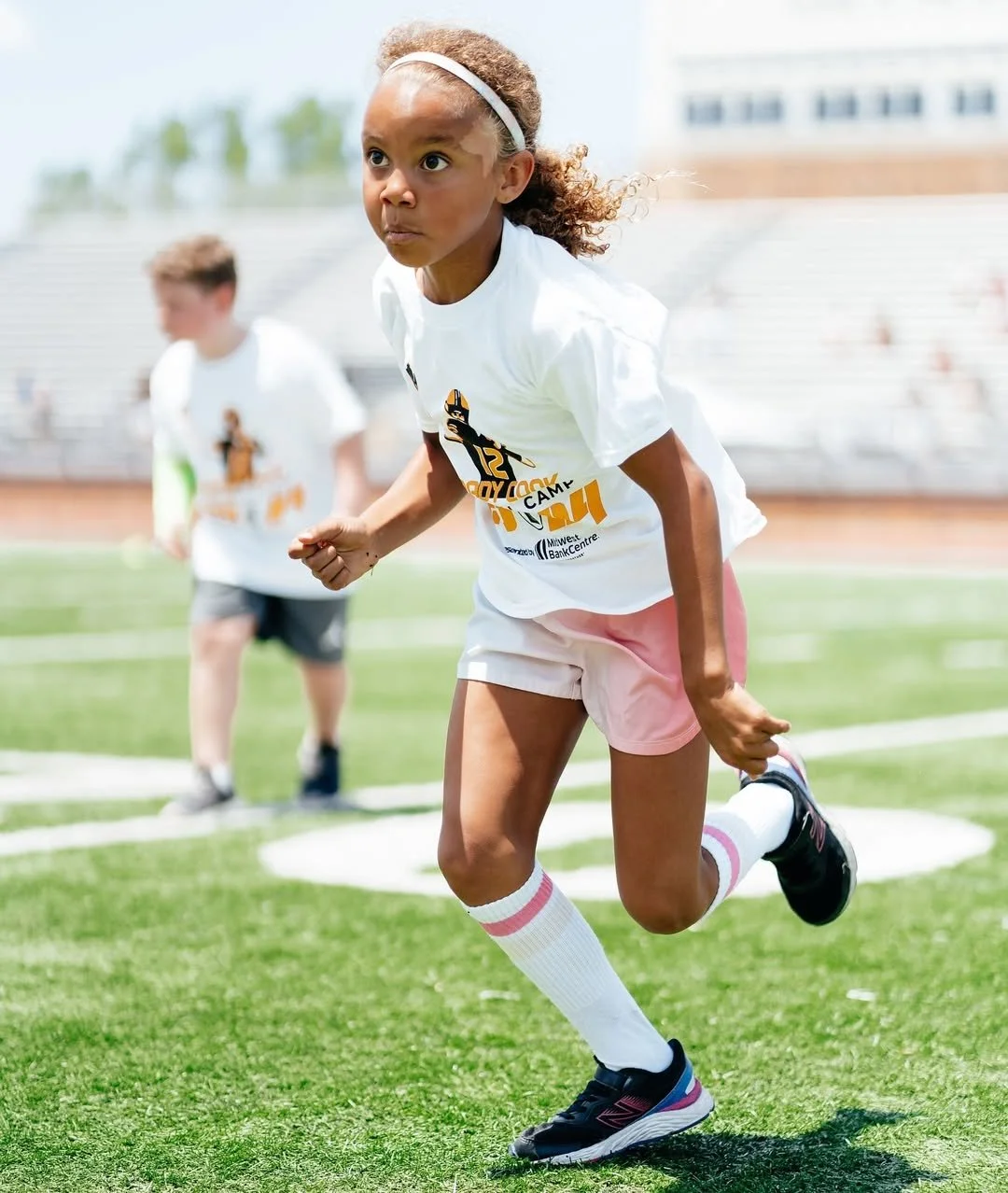 A young girl in athletic clothing running on a sports field during daytime, with a focused expression, other children in the background, and bleachers visible in the distance.