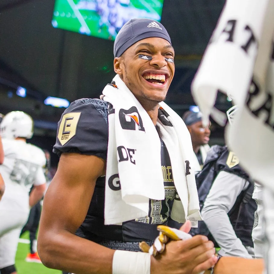 A football player in uniform smiling and holding a beverage, with a towel around his neck, in a locker room or sideline area.