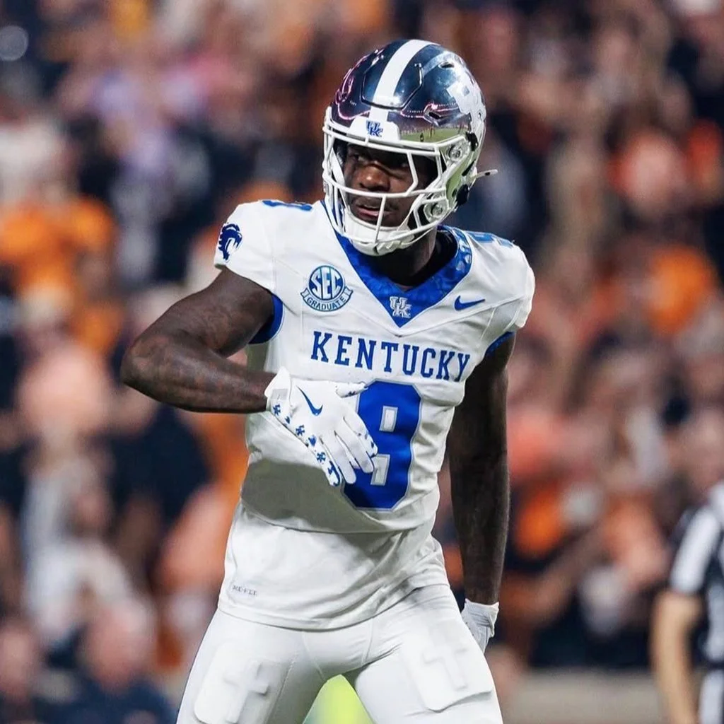 A football player wearing a white and blue Kentucky Wildcats uniform and helmet, standing on the field during a game, with blurred spectators in the background.