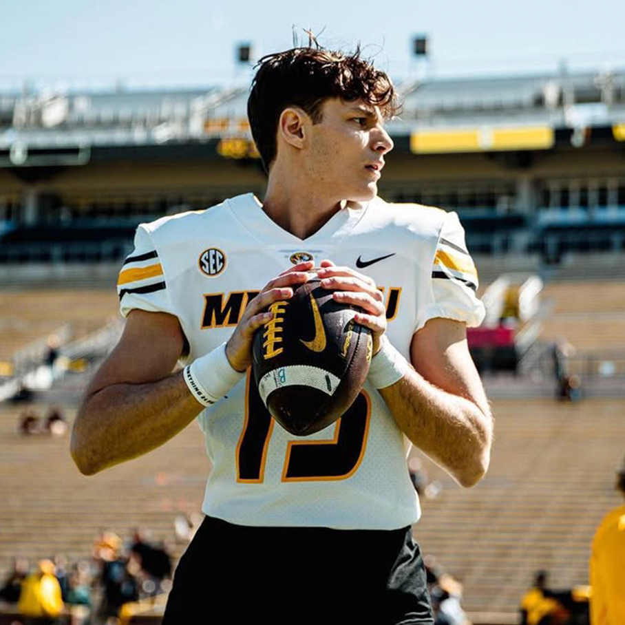 A young male football player in a white uniform with black and yellow accents holding a football on a football field.