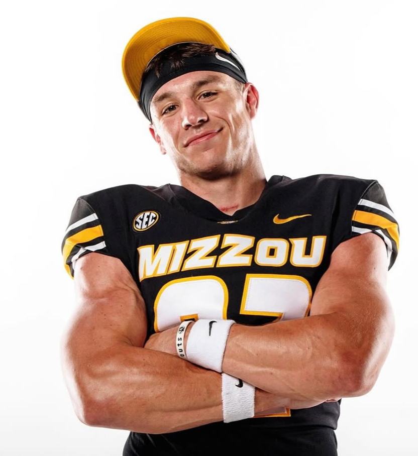 Young man wearing black and yellow football uniform with 'MIZZOU' on it, standing with arms crossed and smiling, wearing a yellow cap and a black headband.