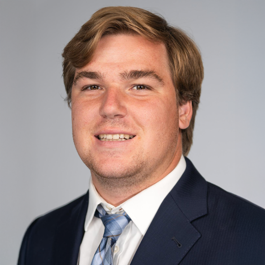 Portrait of a young man with light brown hair, wearing a dark suit, white shirt, and patterned tie, smiling against a plain light gray background.