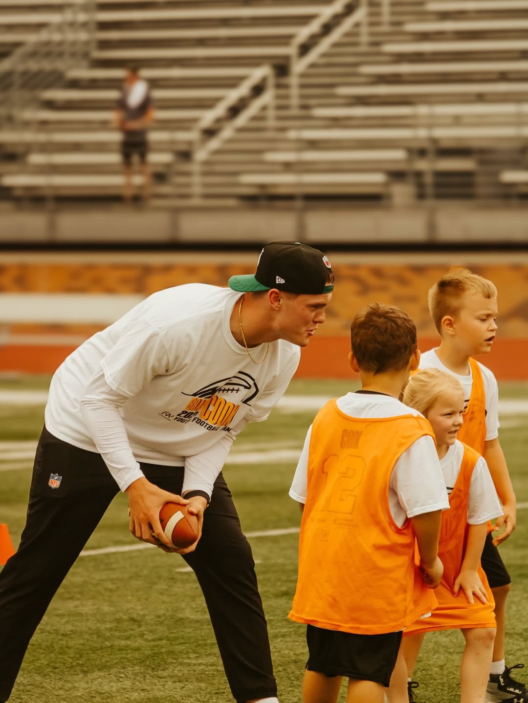 A man coaching children on a football field, holding a football, with children in orange pinnies. Bleachers are in the background.