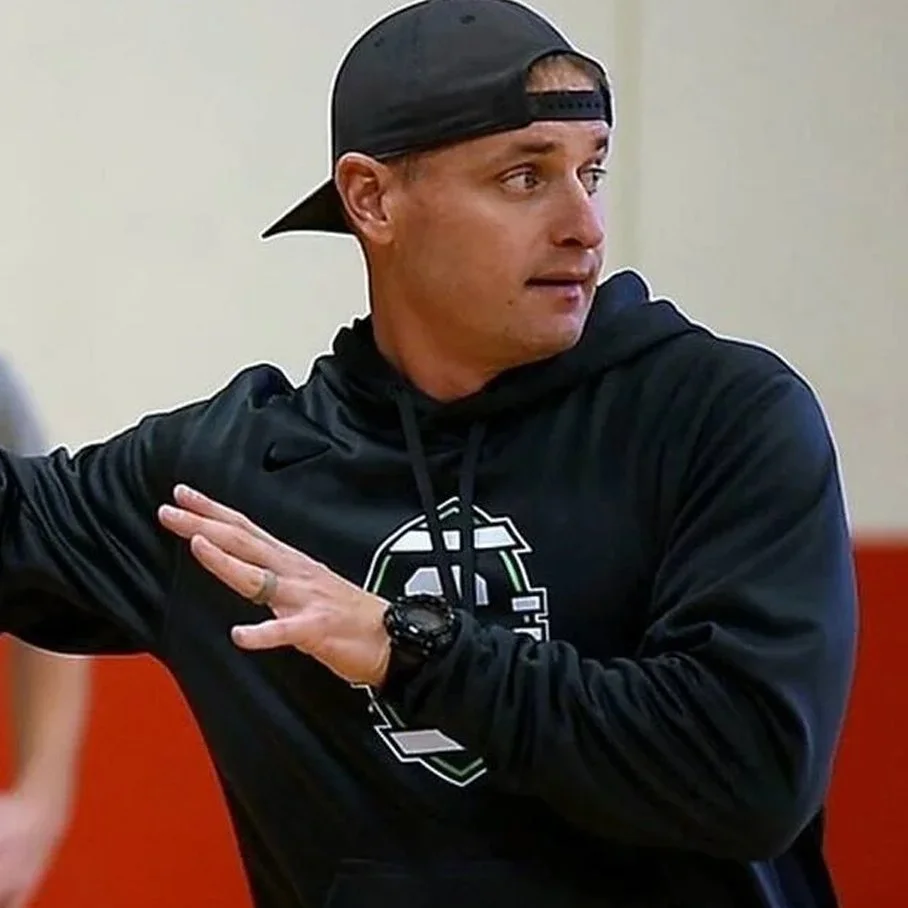 A man wearing a black hoodie and a black backward baseball cap is making a gesture with his arm in a room with beige walls.