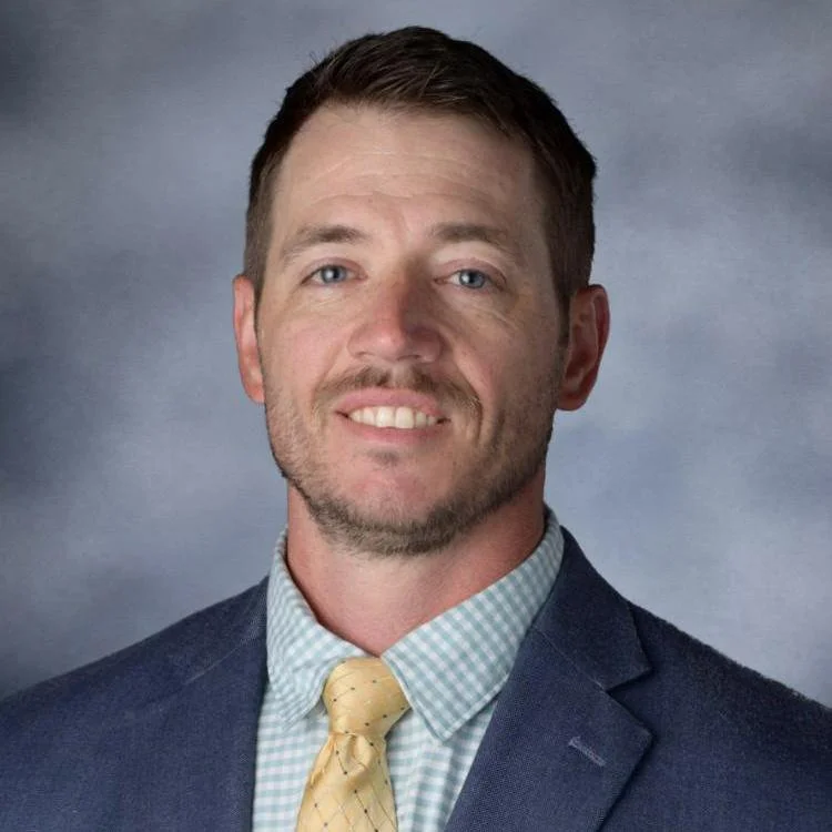 A man wearing a dark blue suit, light blue checkered shirt, and yellow tie, smiling with a neutral gray background.