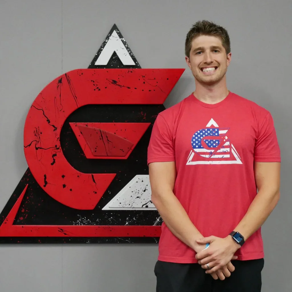 A smiling man wearing a red T-shirt with a patriotic American-themed logo, standing in front of a large black, white, and red logo on a gray wall.