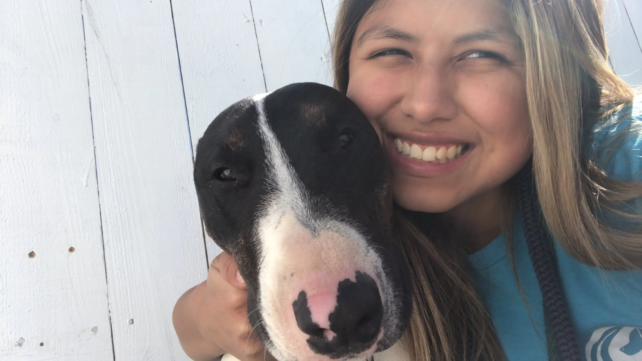 A woman smiling as she holds a black and white dog close to her face, in front of a white wooden wall.