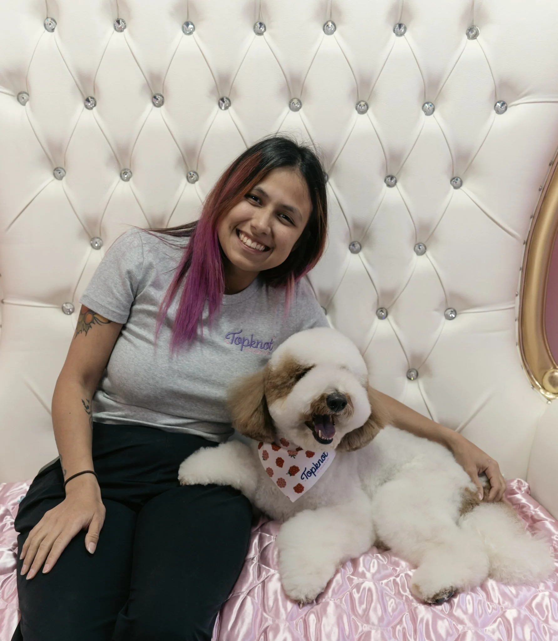 Alisa sitting on a pink satin chair, with a dog wearing a 'Topknot' bandana, in front of a tufted white headboard with crystal accents at Topknot Pet Salon & Spa in Los Angeles, California